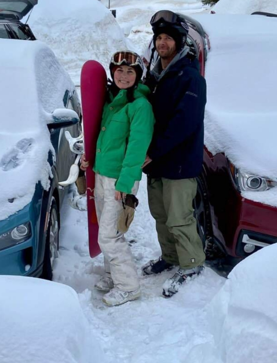 A woman in a green jacket, ski helmet, ski goggles, snowboard boots carries a snowboard, stands next to a man wearing a ski helmet, ski goggles, a winter jacket and ski boots. They stand between two parked cars covered in snow.  
