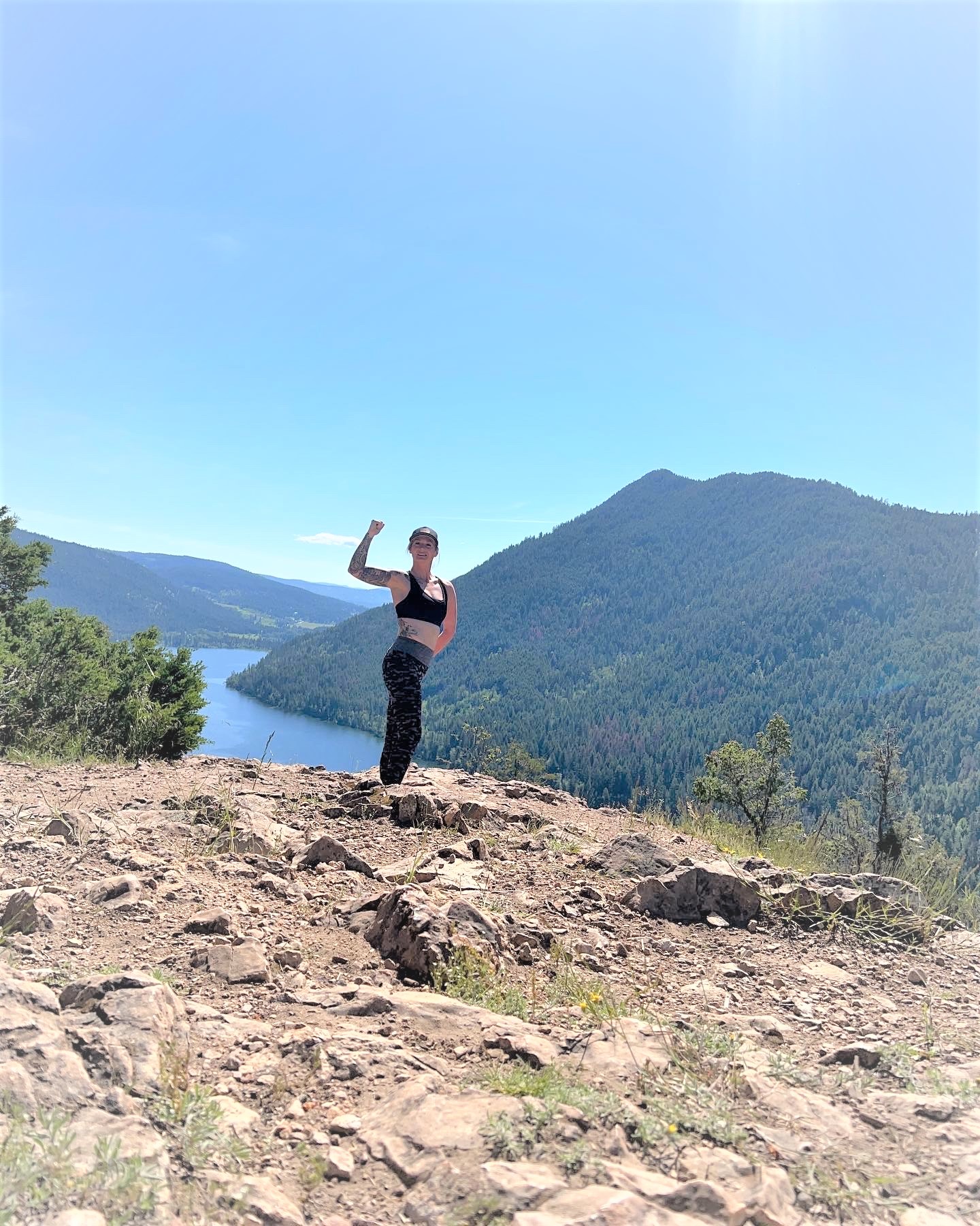 A woman in outdoor active wear flexes her right arm while standing on a rocky cliff. There are large hills with trees and a body of water in the background.
