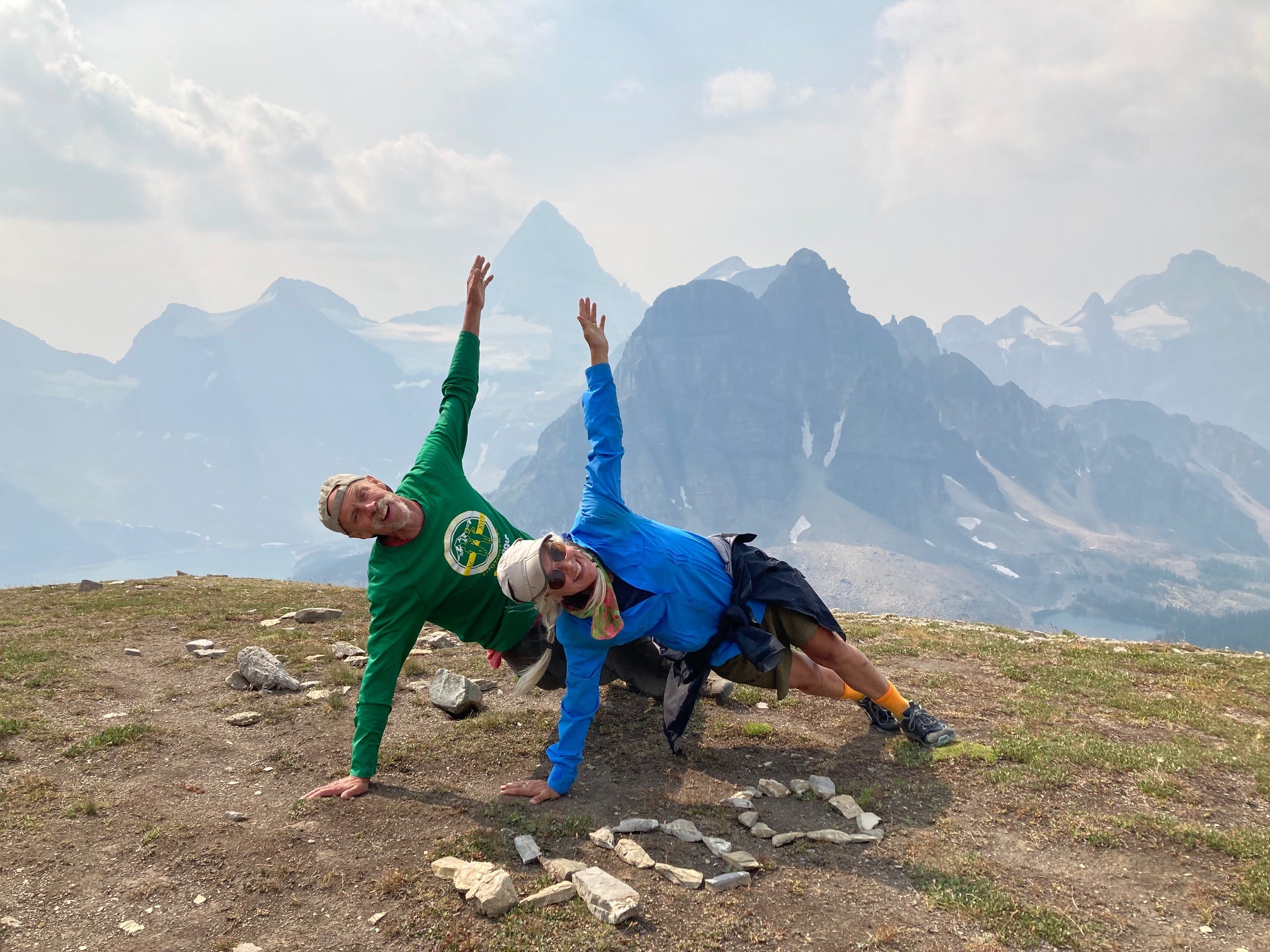 Man and woman on mountain top wearing ball caps and hiking gear, doing side push ups.