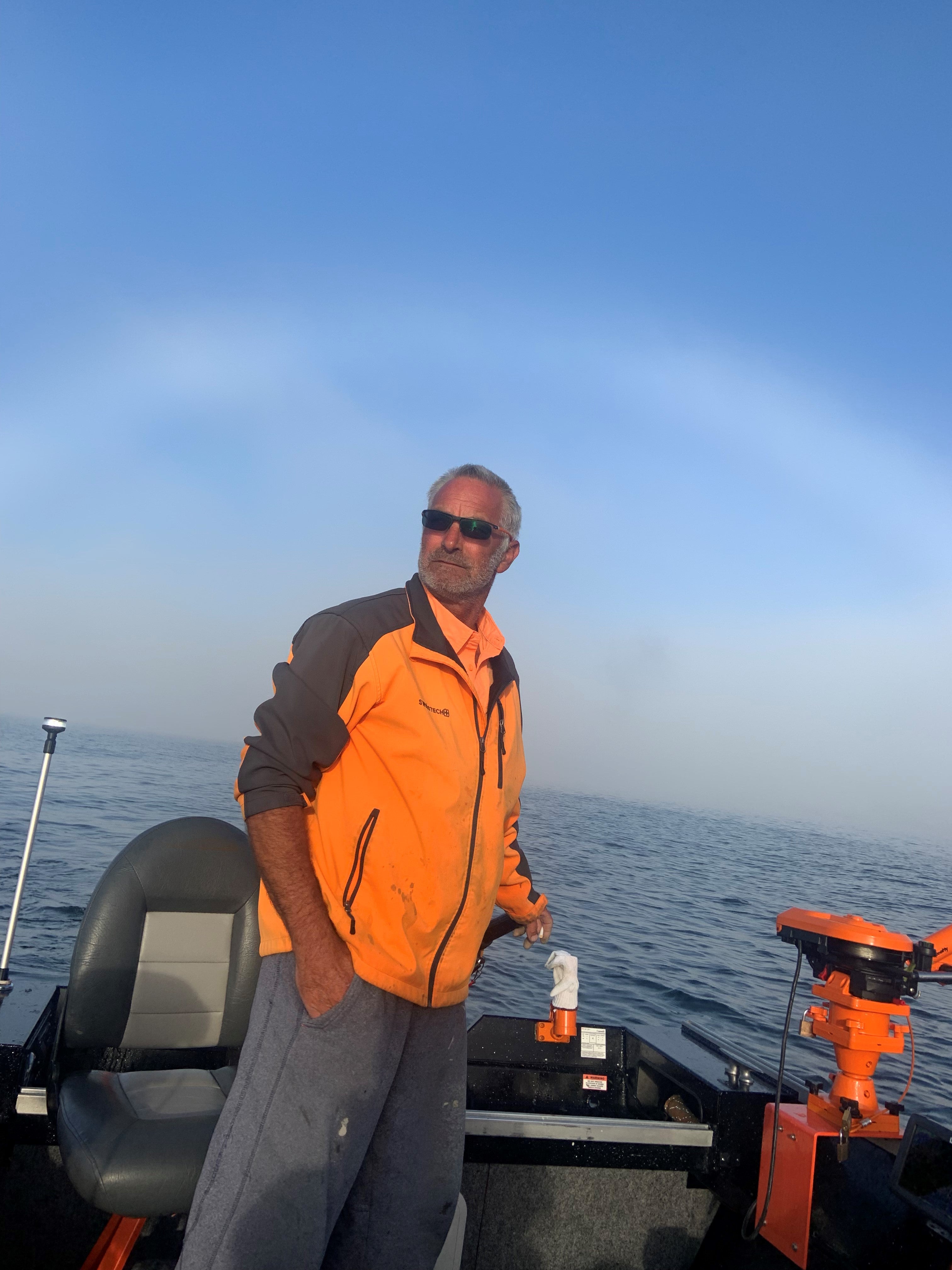 A man in an orange and grey jacket steering a boat on a body of water.