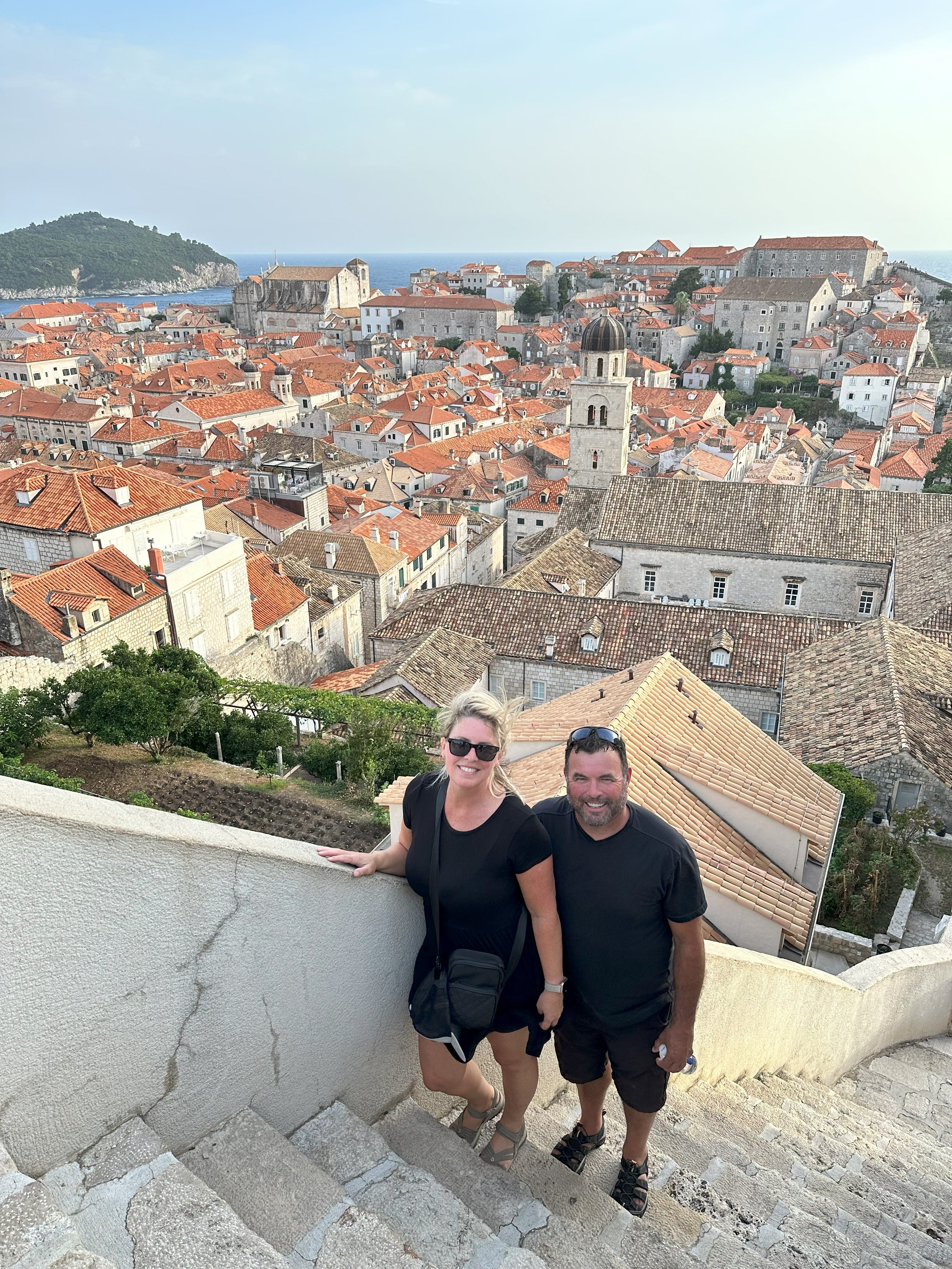 A couple, blonde woman wearing sunglasses, black top and shorts and beige sandals, and a man wearing black top, shorts and sandals, stand on stairs next to a wall overlooking houses