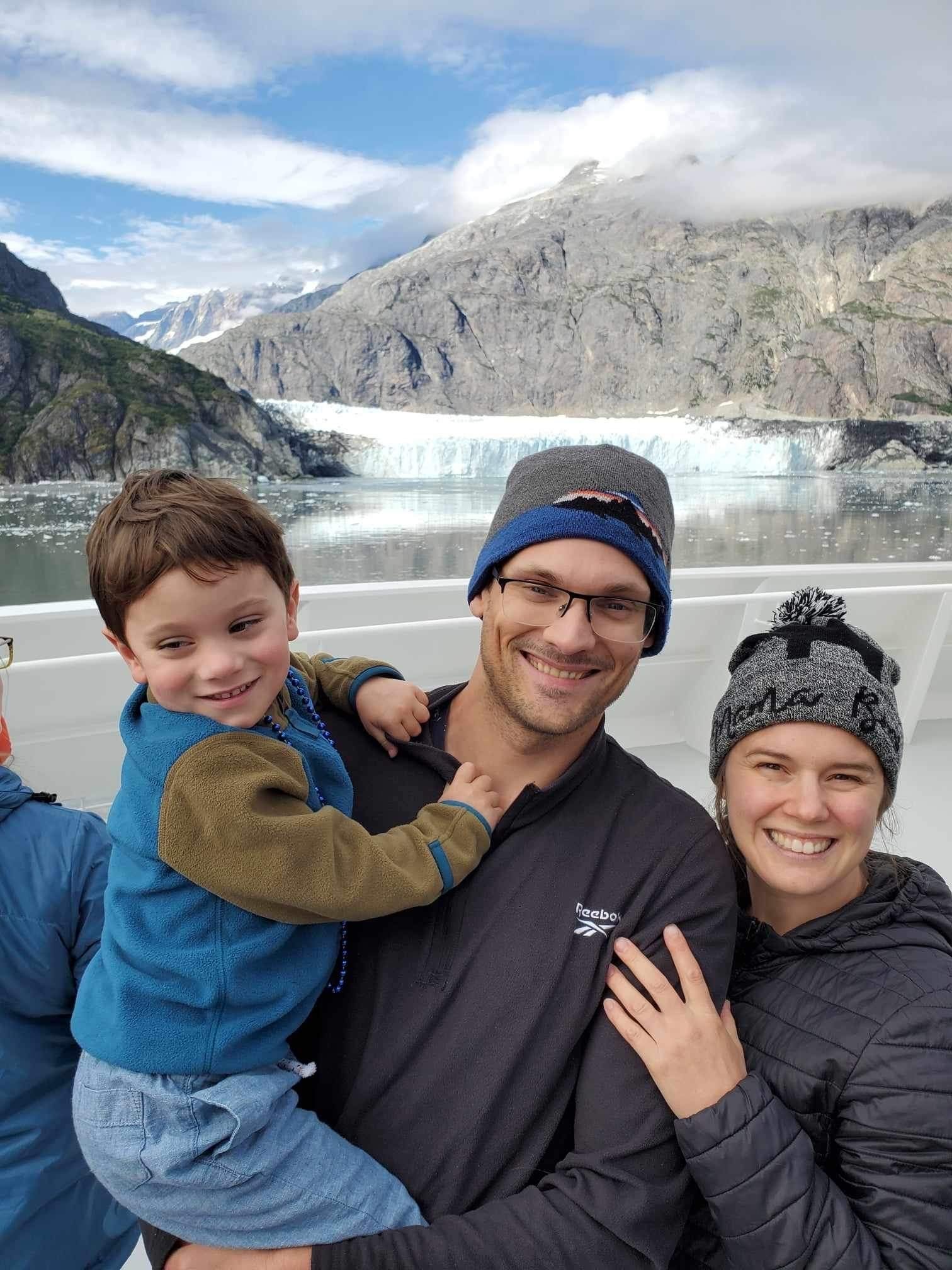 A smiling man, woman and child, all wearing winter gear, pose for a picture outside with water and mountains in the background.