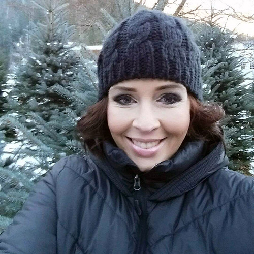 The face of a woman in a toque, in front of evergreen trees.