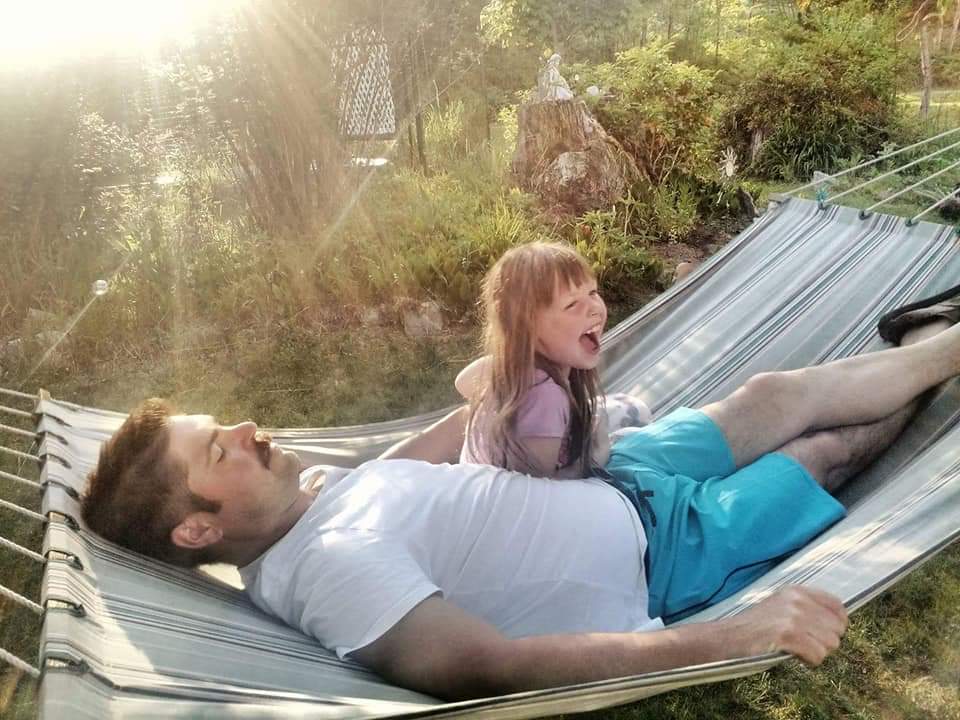 A man in t-shirt and shorts lays in a hammock in a green yard, with a young girl in a purple outfit sitting up next to him, appearing to be laughing.