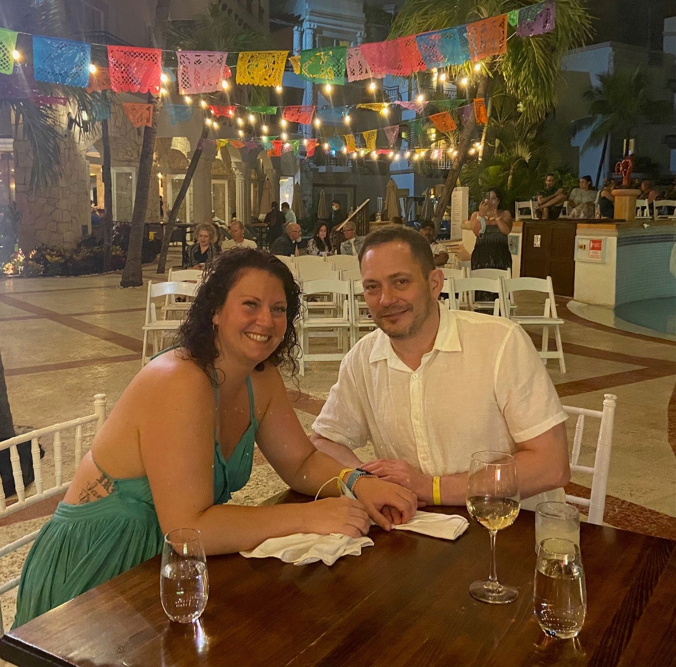 A woman on the left in a blue sundress and a man on the right in a white linen shirt seated at a table with drinks on it, with a background of people sitting in white chairs in rows and coloured flags hanging from strings above.