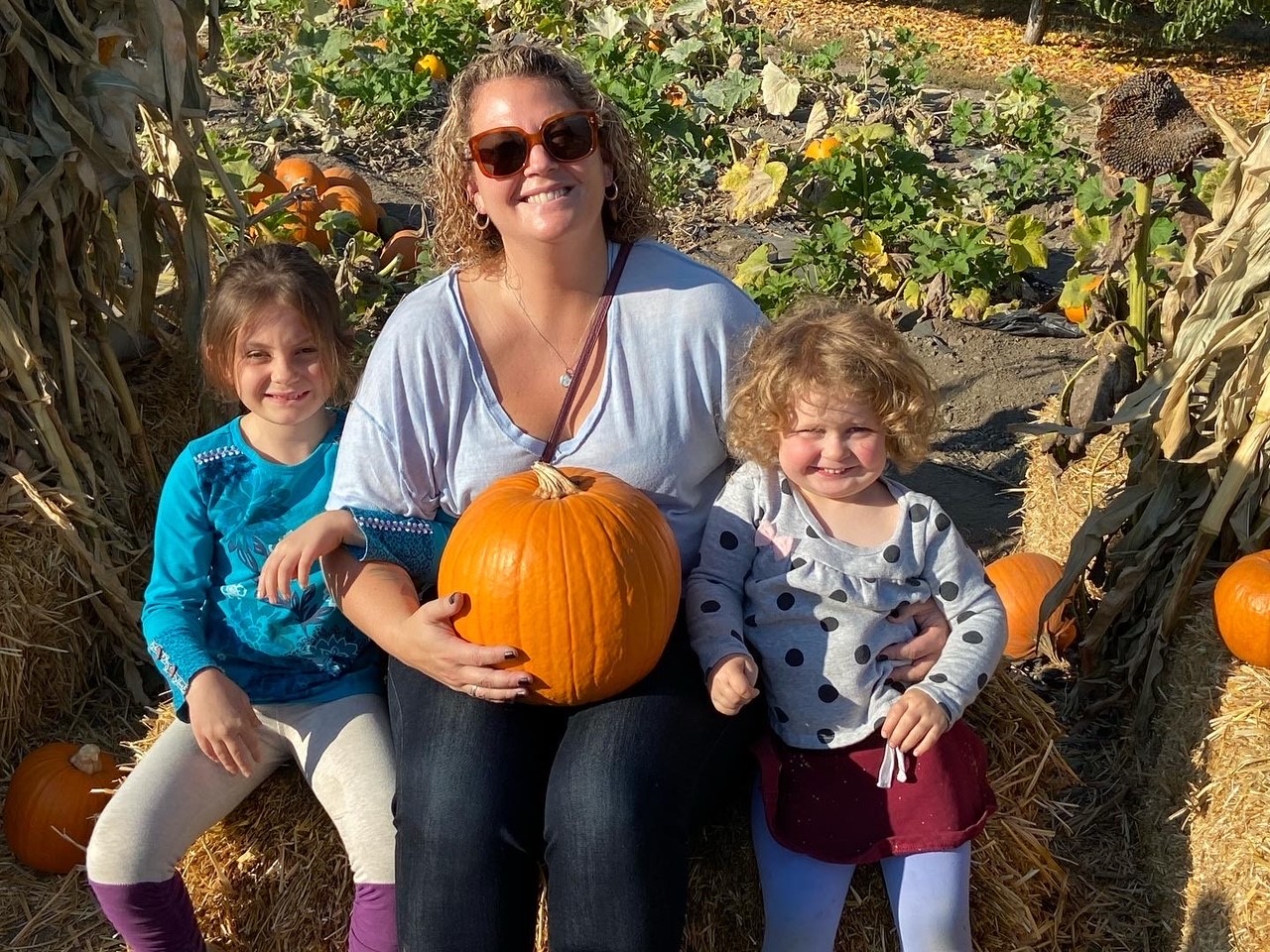 A woman with wavy blonde hair, wearing sunglasses, with a light blue t-shirt and jeans, holding a pumpkin, sitting on a hay bale between two children, the one on the left with a bright blue sweatshirt and grey and purple leggings, and on the right with a grey and black polka dot sweatshirt, red skirt and blue leggings, with a pumpkin patch in the background.