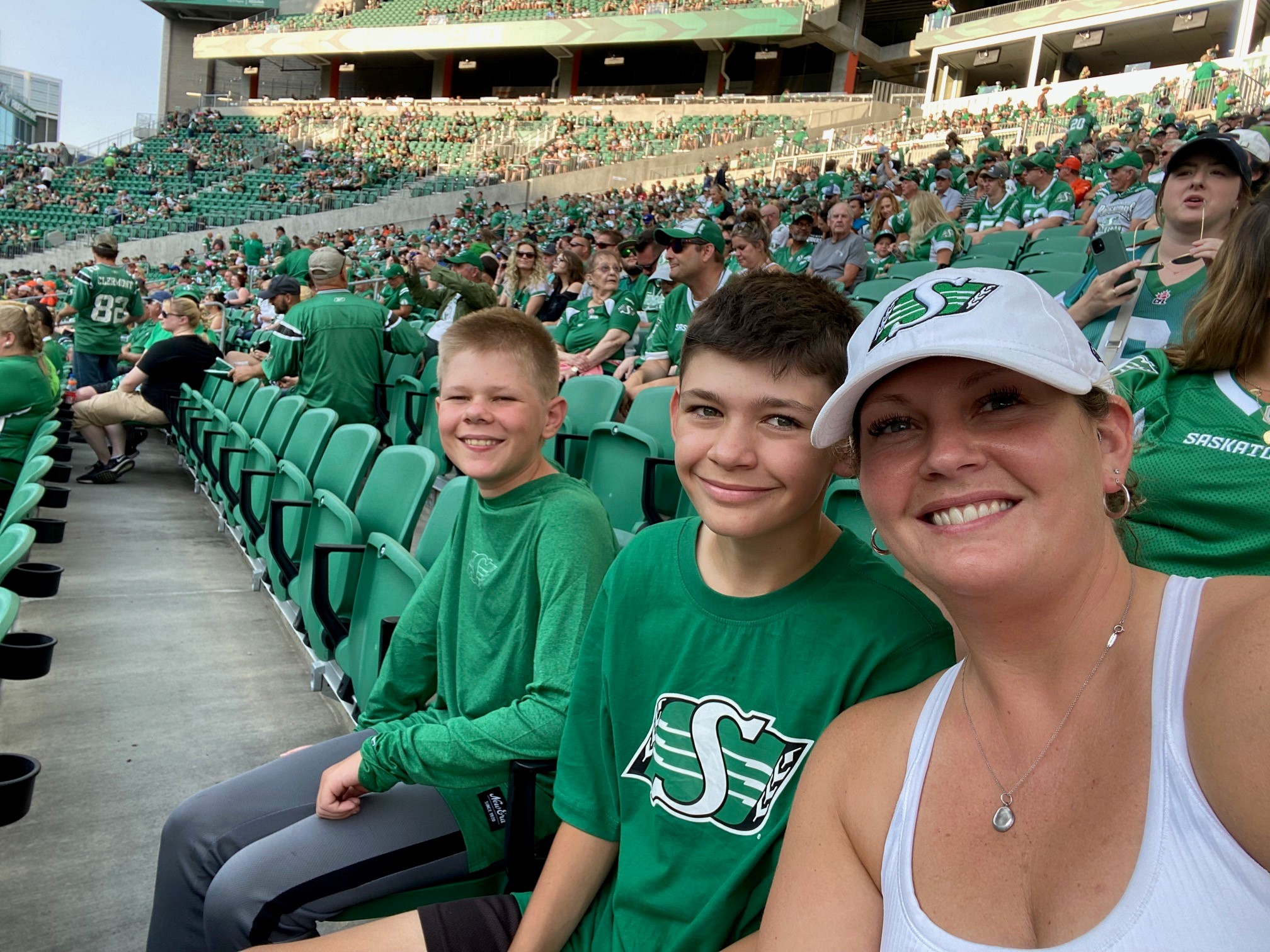 A women on the right with a white Saskatchewan Roughriders ballcap and white tank top, with two boys to the left, wearing green Roughriders shirts, in a stadium with green seats and many people wearing Roughriders merchandise in the background.