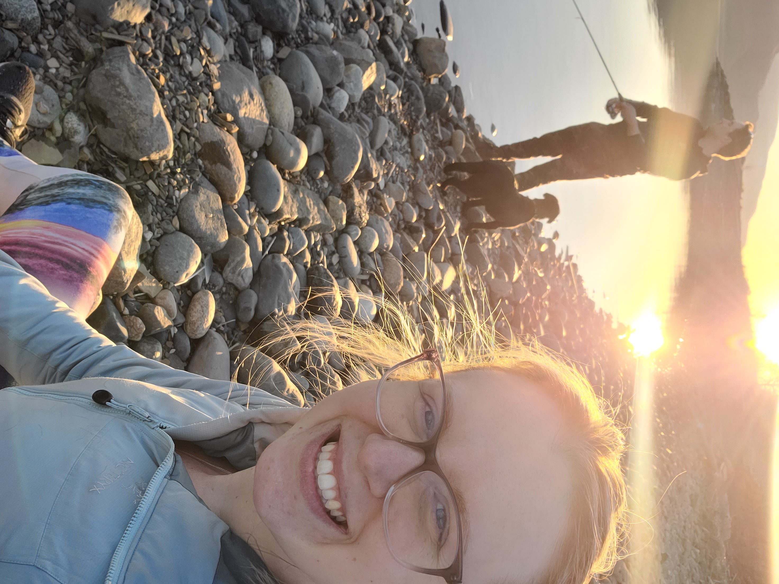 Woman wearing glasses at rocky beach and man with dog at water’s edge with sunset.