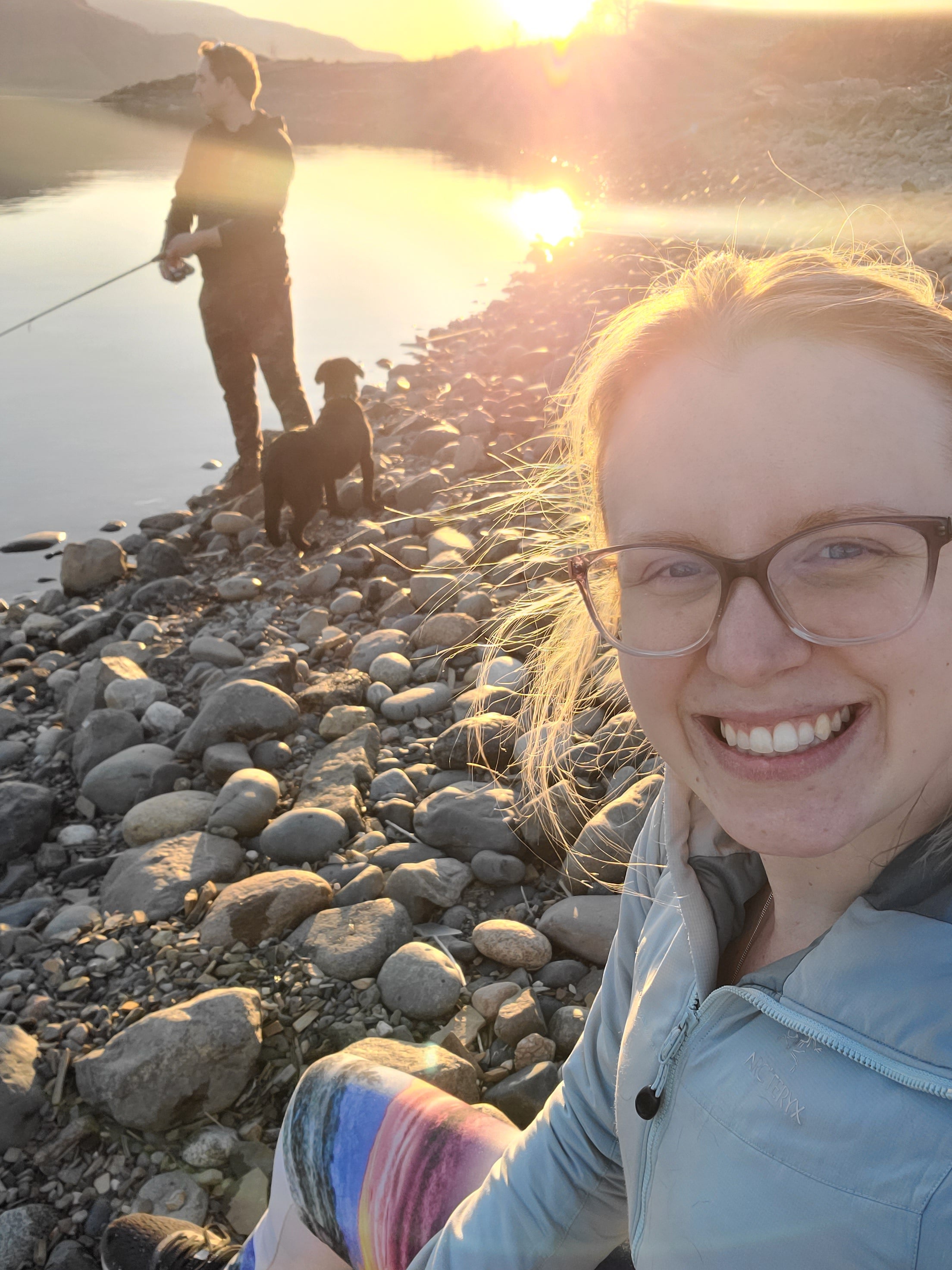 Woman wearing glasses at rocky beach and man with dog at water’s edge with sunset.