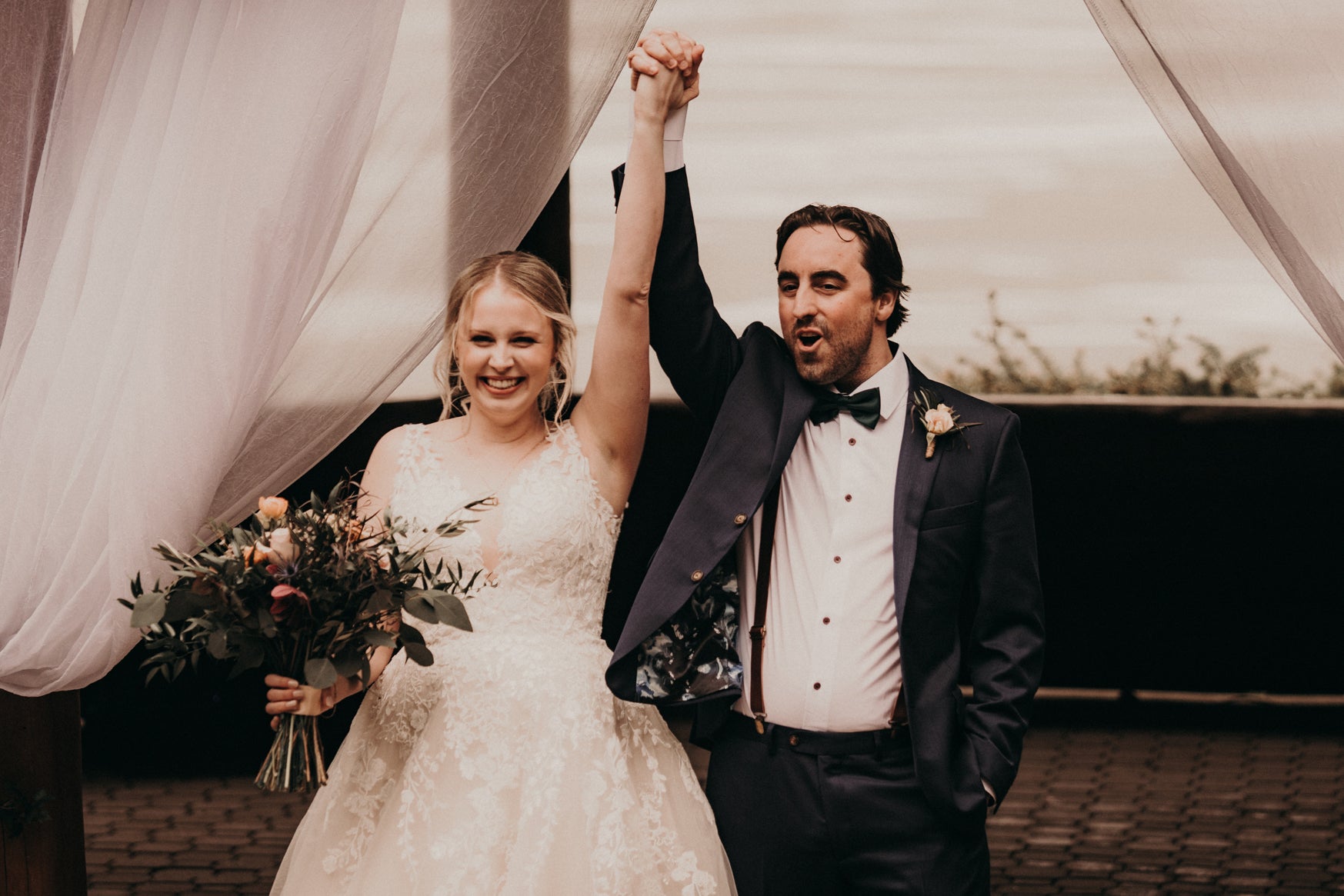 Smiling bride carrying bouquet of flowers in one hand while holding groom’s hand in the air.