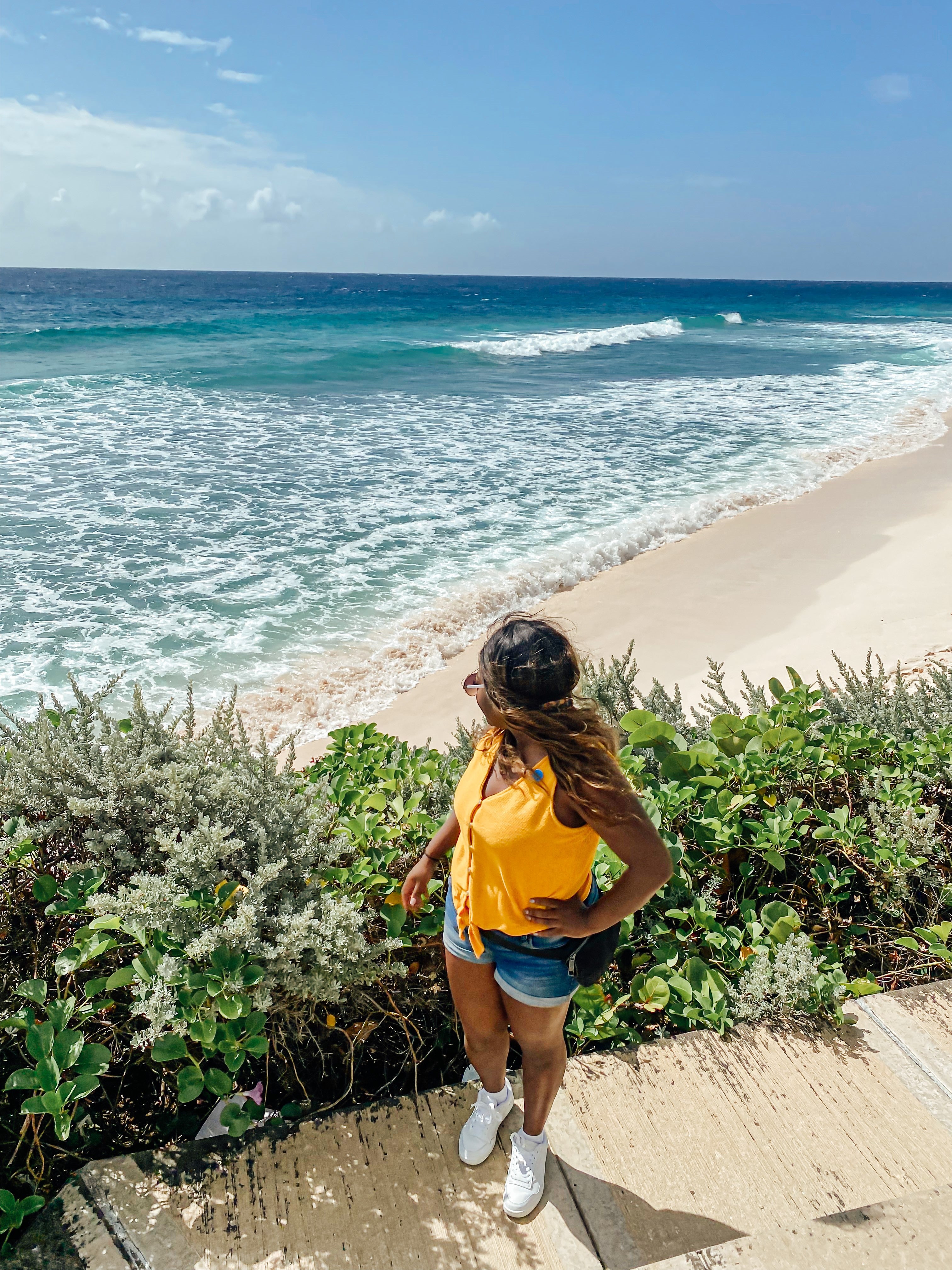 A woman wearing summery clothing stands on a tropical beach by some shrubbery and looks out at the ocean.