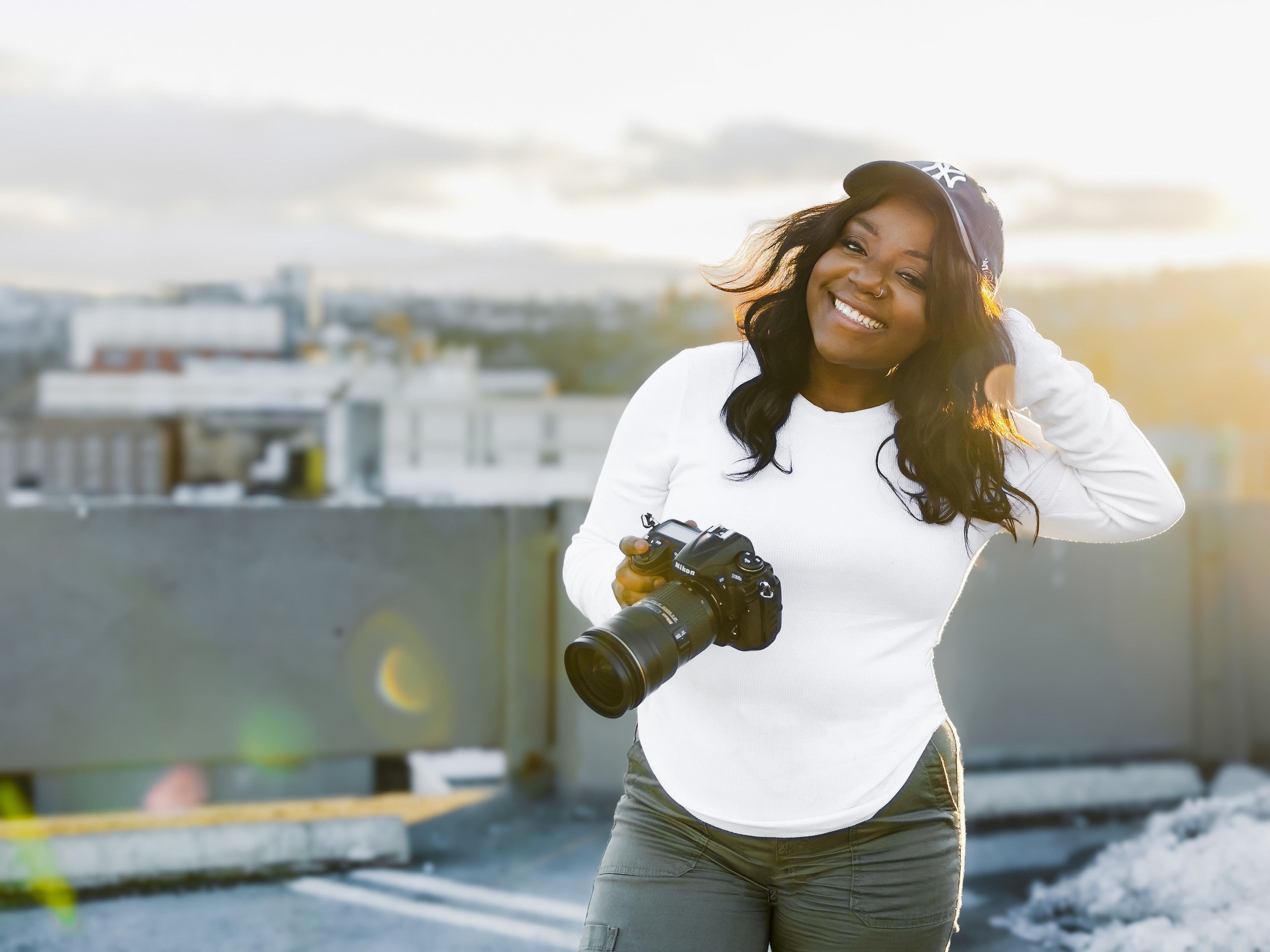A smiling woman with long hair wearing casual clothes and a New York Yankees hat holds a camera in one hand while standing outside. The scene in the background is blurred.