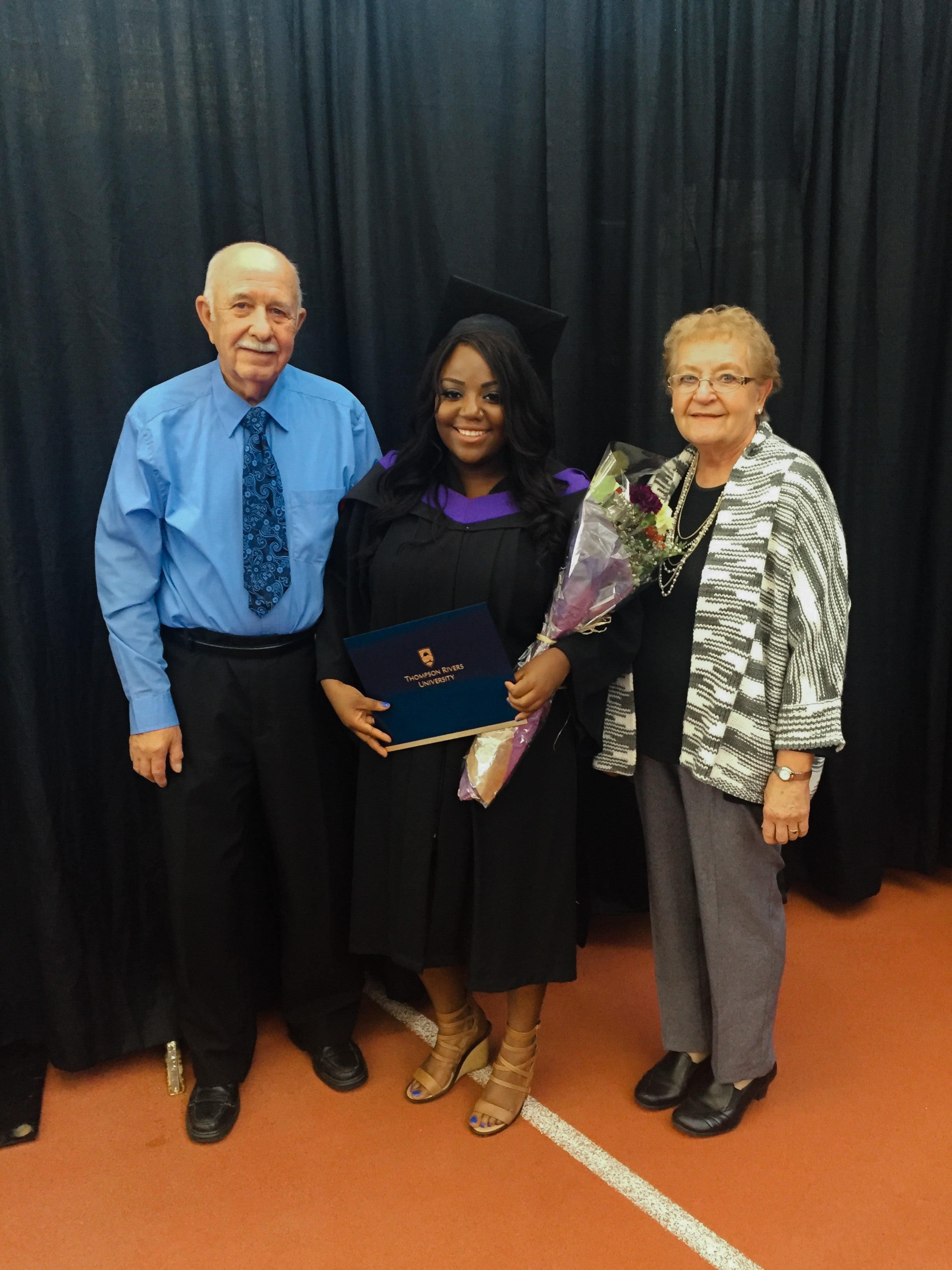 A smiling woman dressed in a graduation outfit and holding flowers and a diploma poses for a picture with two older adults on either side of her. They are standing on a gymnasium floor with black curtains as a backdrop.