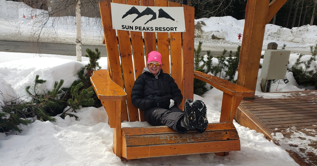 A woman in winter gear sits outside on a big wooden chair with a black Sun Peaks Resort logo on it.