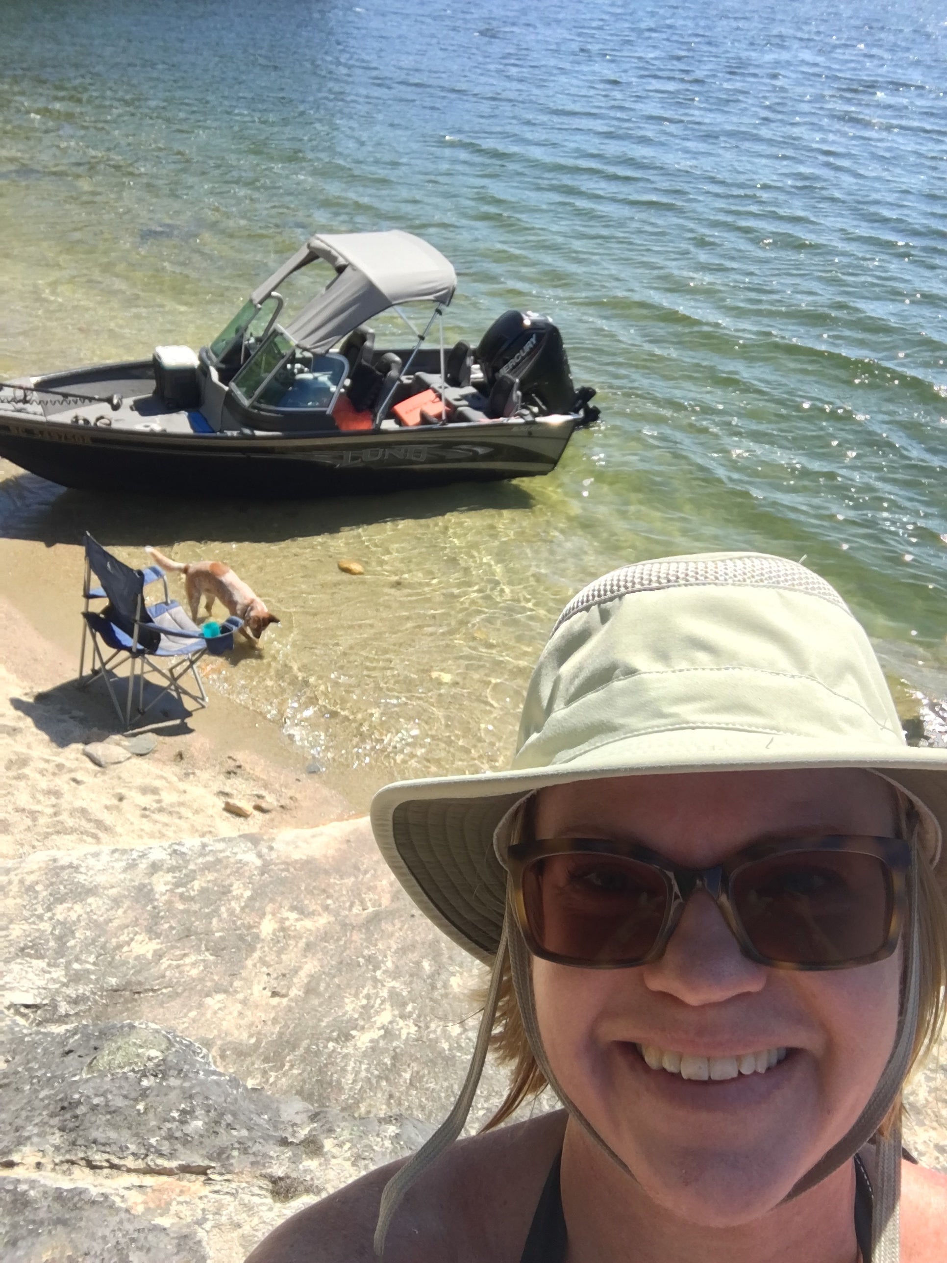 A woman takes a selfie near a lake with a dog and boat in the background