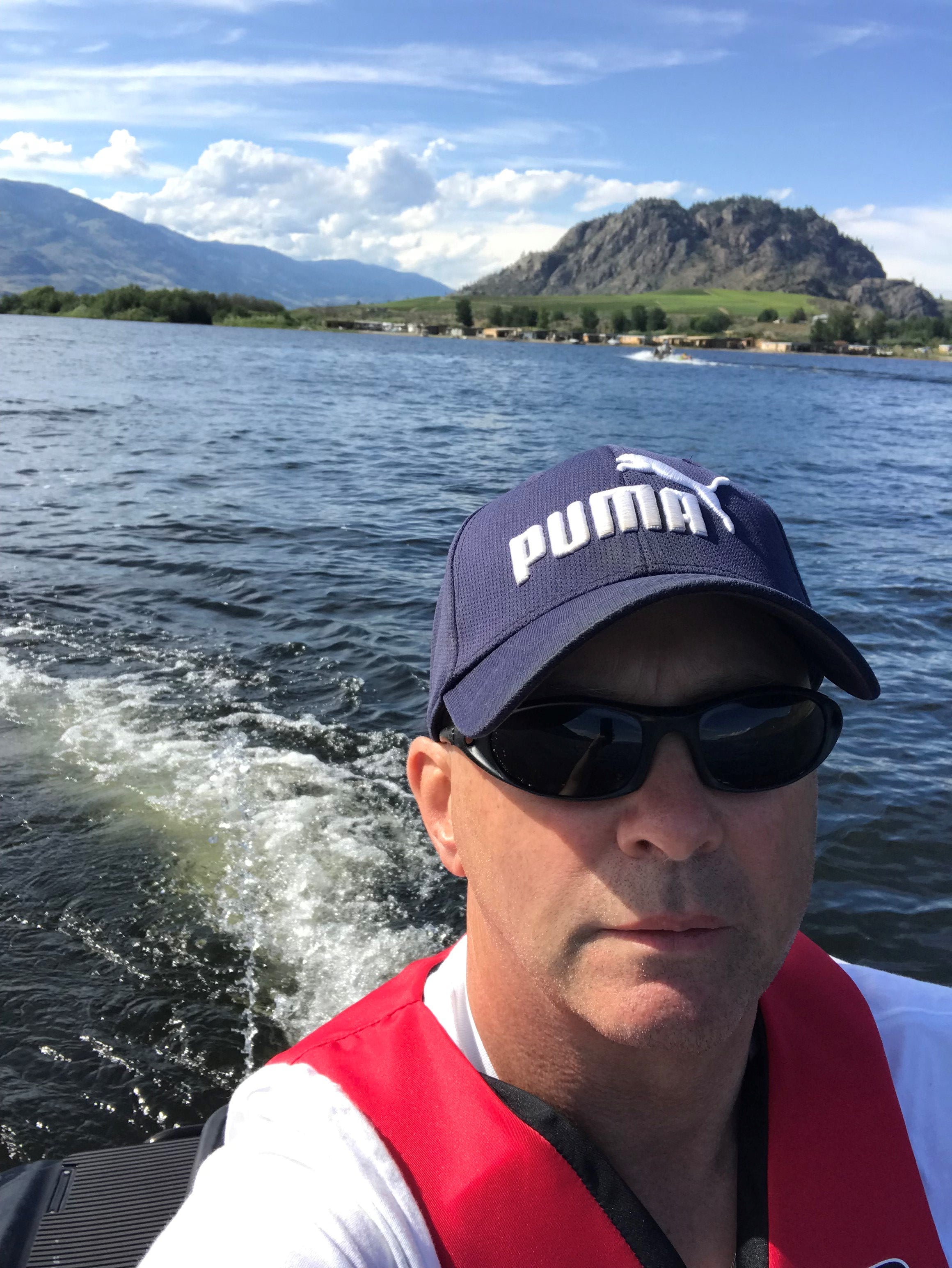 A person in a navy blue ballcap with Puma logo, in red life vest on a boat in the water with land with green grass and a small mountain, and blue skies.