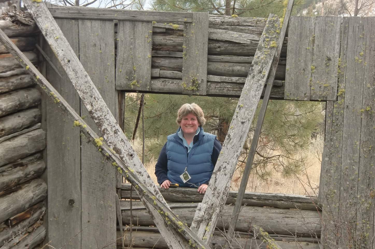 A woman standing in the forest poses for a picture while looking through an opening in a wooden wall.