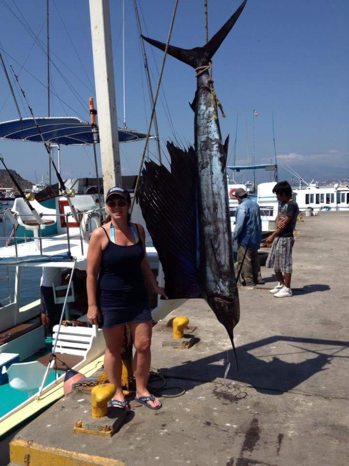 A woman on a pier wearing a dark ball cap, camisole and shorts standing to the left of a large dangling fish, in front of a boat, with two people in the background to the right of the fish.