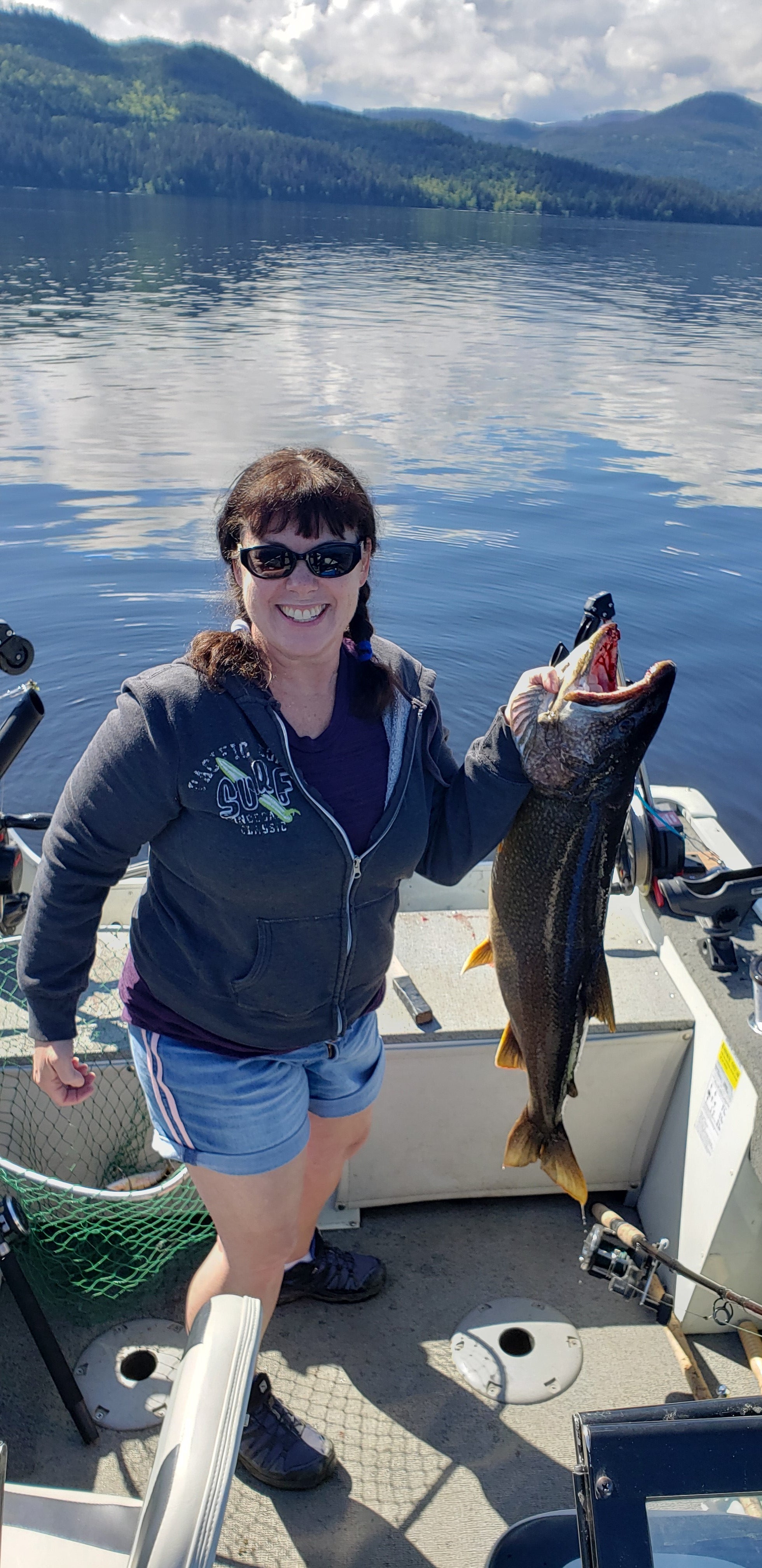 A woman with dark brown hair in two braids, wearing a grey hooded sweater and blue shorts, holding a fish while standing in a boat on a lake with trees and hills in the background.