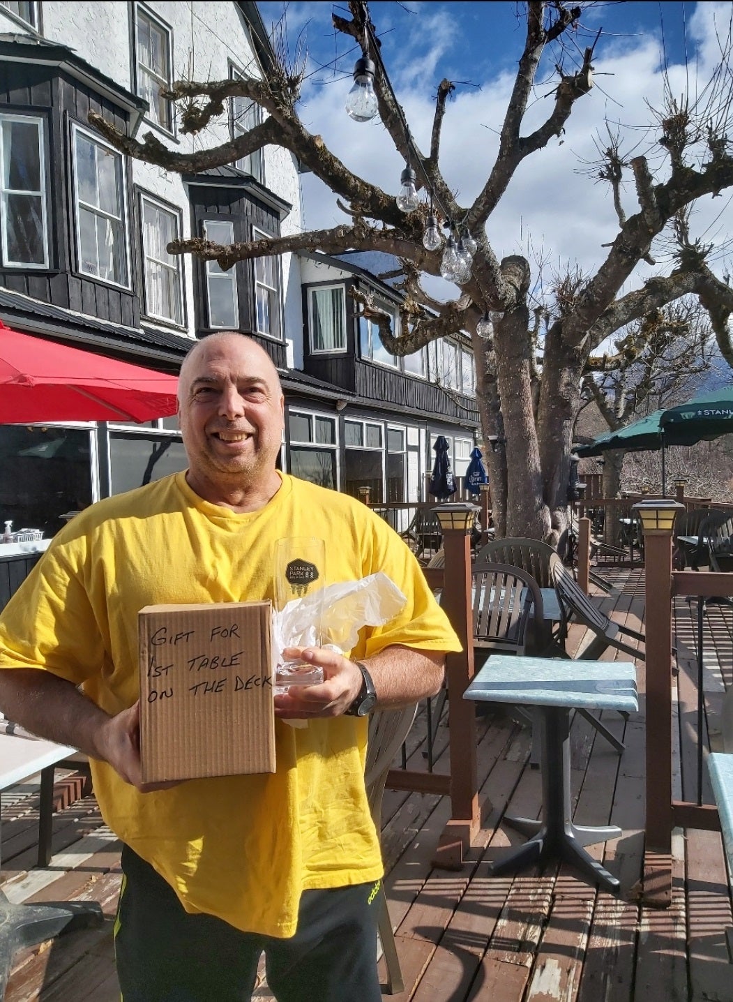 A person standing on a patio outside a large complex holds a box that says “Gift for 1st table on the deck.”