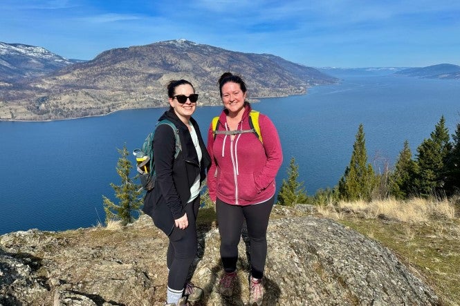 Two people in leggings and sweatshirts standing on a rock with a lake and mountains in the background.