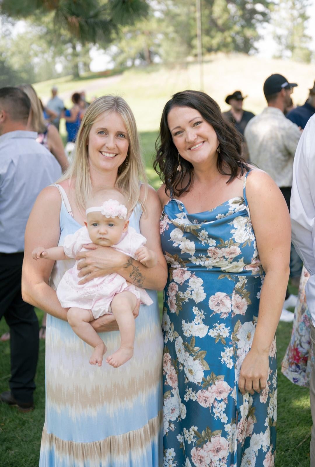 Two people standing outdoors wearing long summer dresses holding a baby.