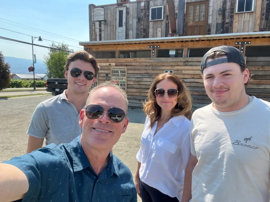 Woman, man, and two boys in sunglasses standing in sunshine in front of older wooden building.
