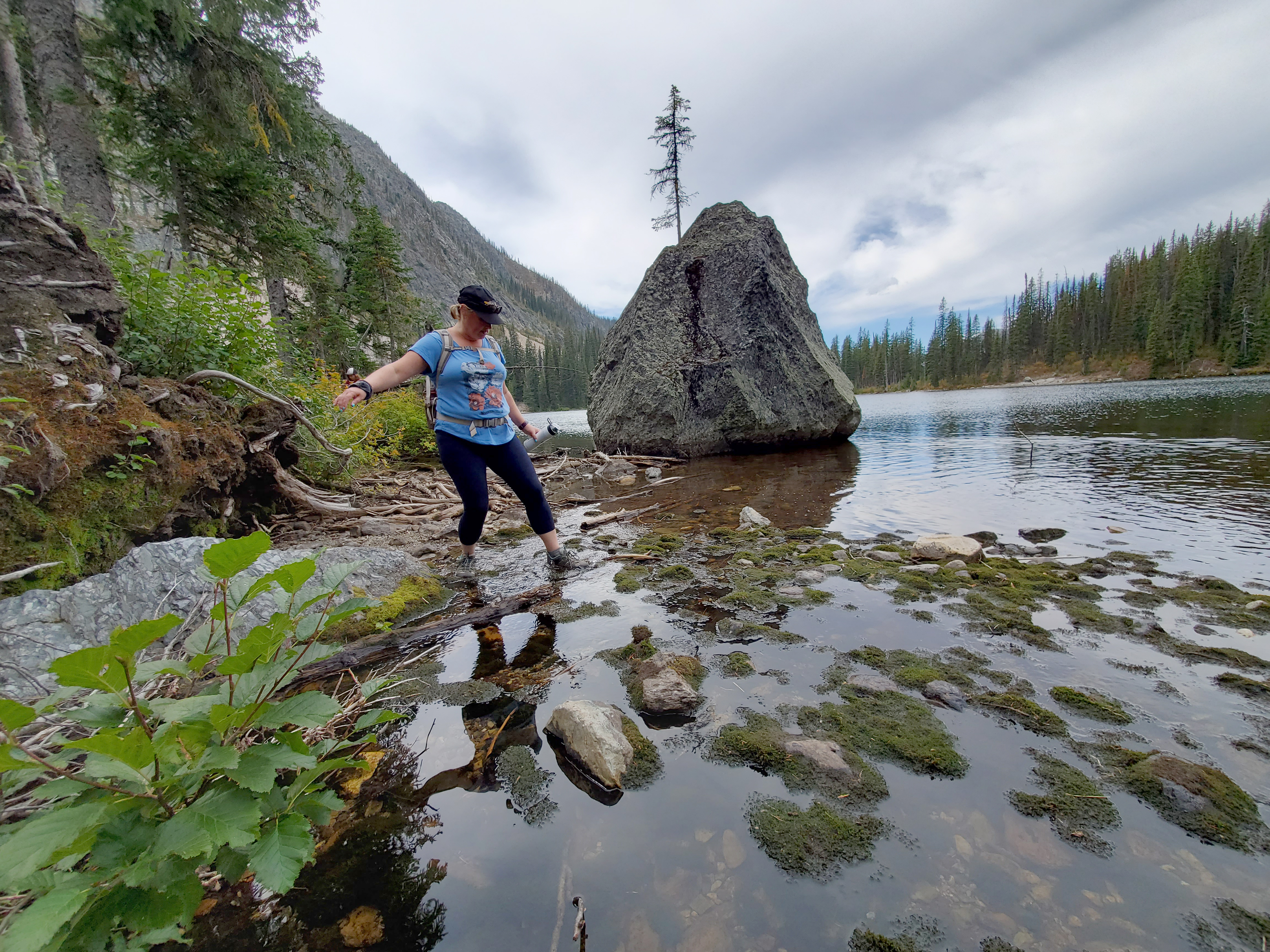Hiking woman wearing blue t-shirt and ball cap, stepping into water at edge of lake with rocks and moss, and with trees and mountain in background