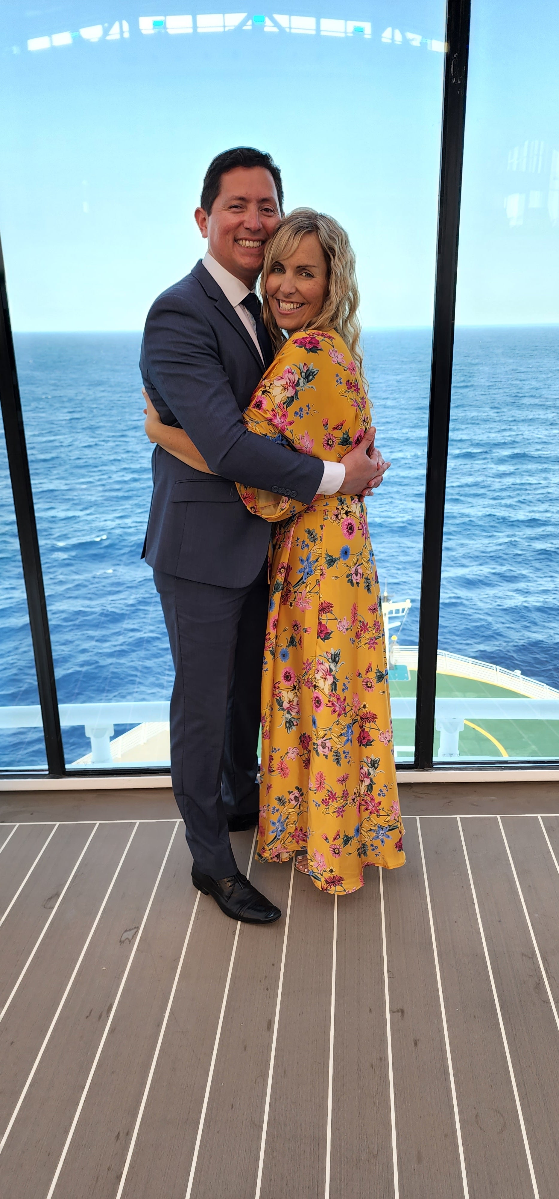 A man and woman in formal evening wear pose for a picture on a cruise ship by a window with the ocean in the background.