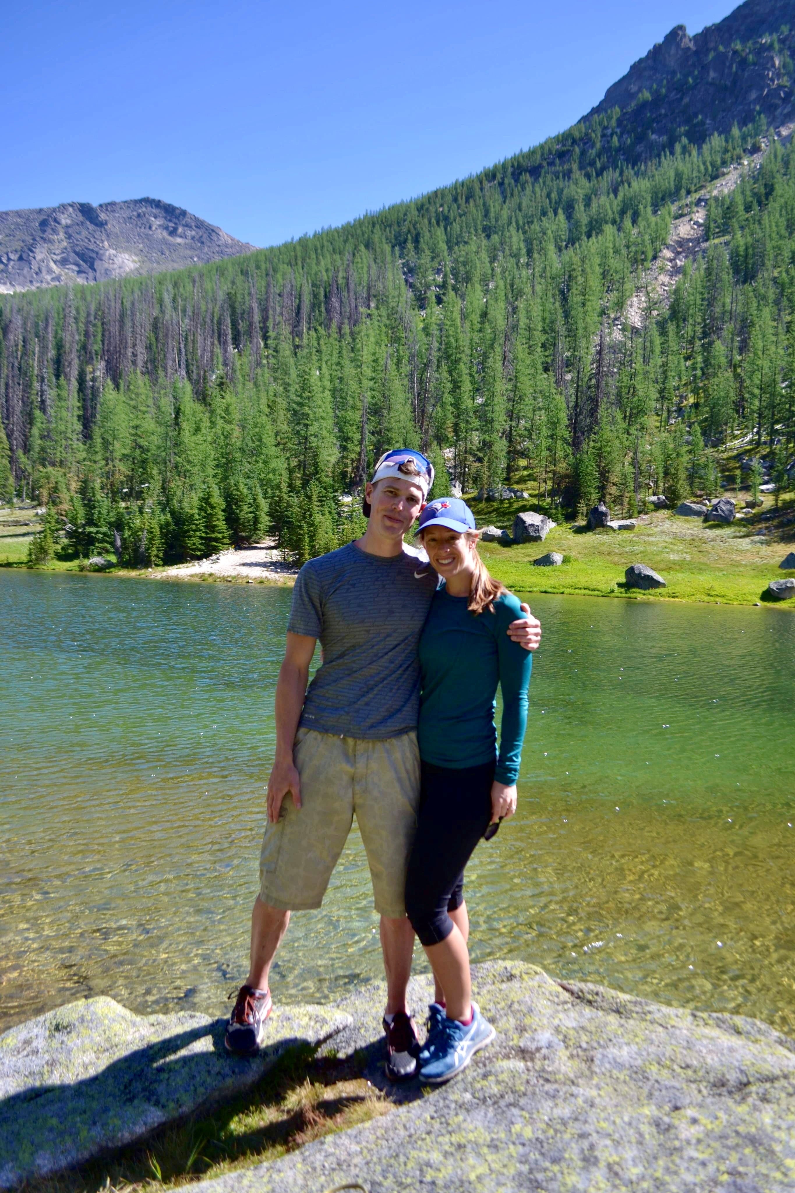 Two people with an arm around each other pose for a picture while standing on a rock in front of clear water and a tree-covered hill