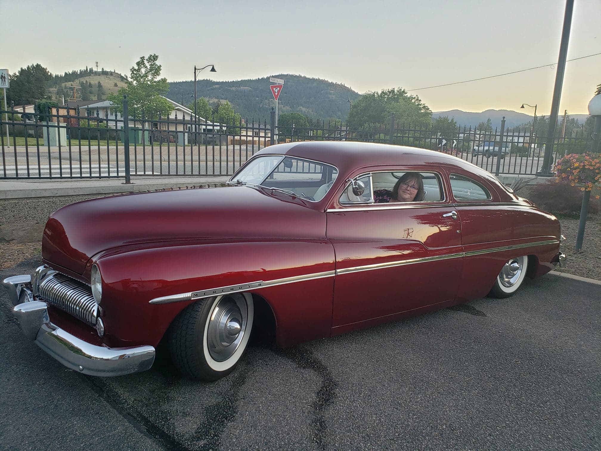 Woman inside burgundy 1949 Mercury hot rod with park and mountain in background.