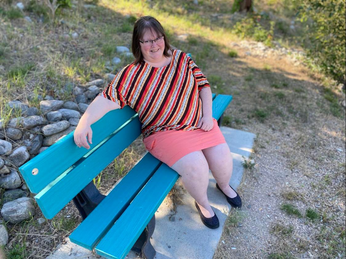 Woman wearing striped top, pink skirt and black shoes, with short brown hair and glasses, sits on blue park bench.