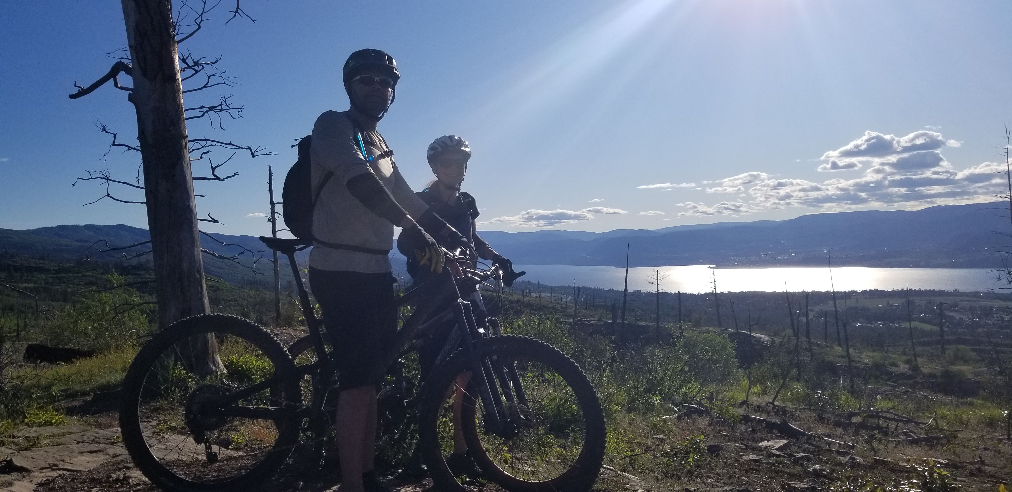 A man and woman on bikes on a hill with a lake behind.