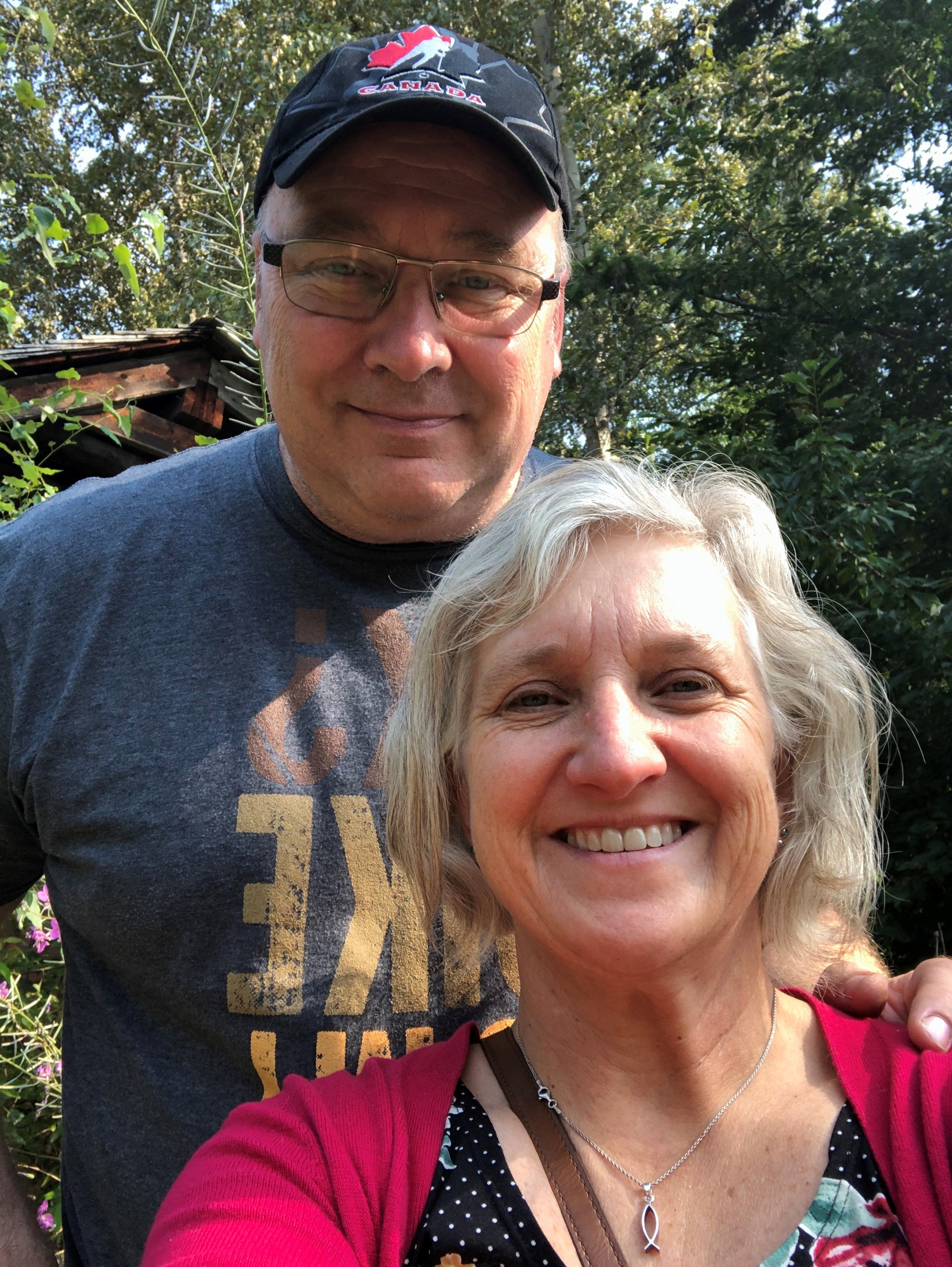 A man, wearing a ballcap, and a woman with grey hair standing in a forest.