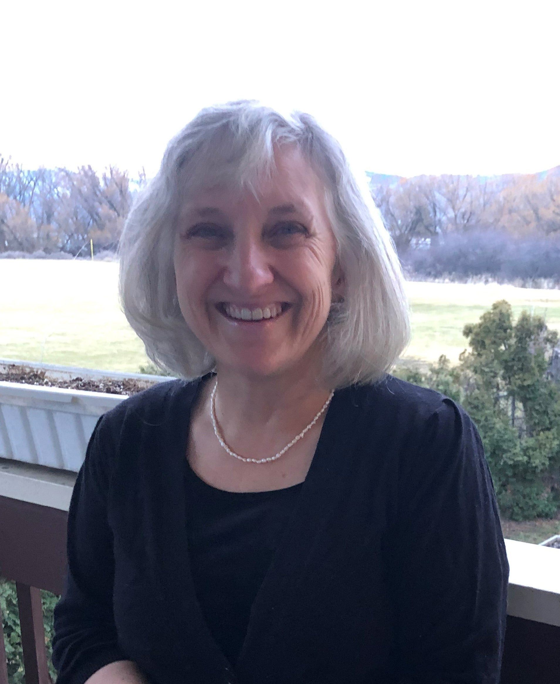 A woman with grey hair in a black outfit wearing a pearl necklace.