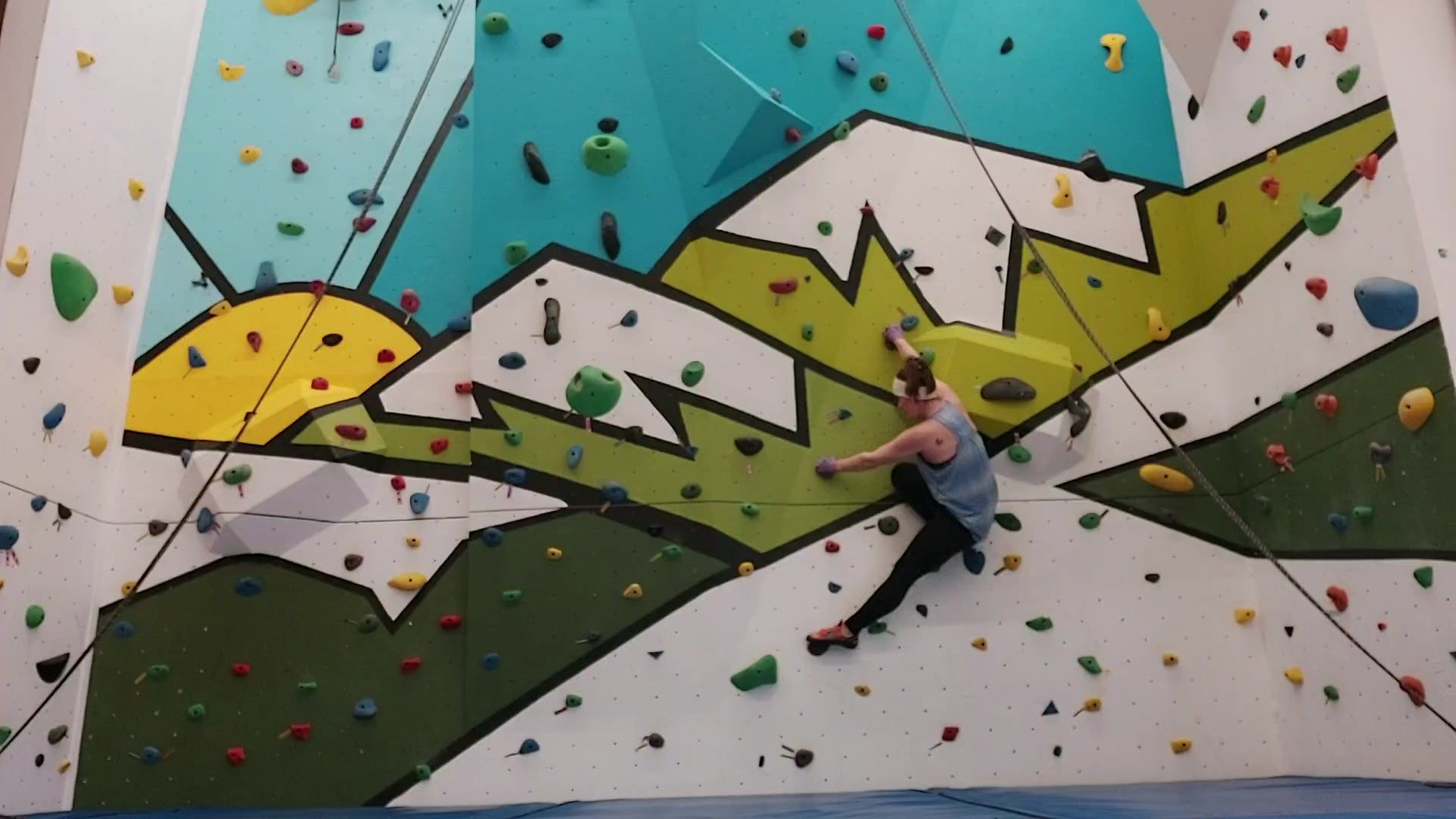 Person wearing teal tank top and black pants balances across pegs on indoor rock-climbing wall with painting of sun and mountains.