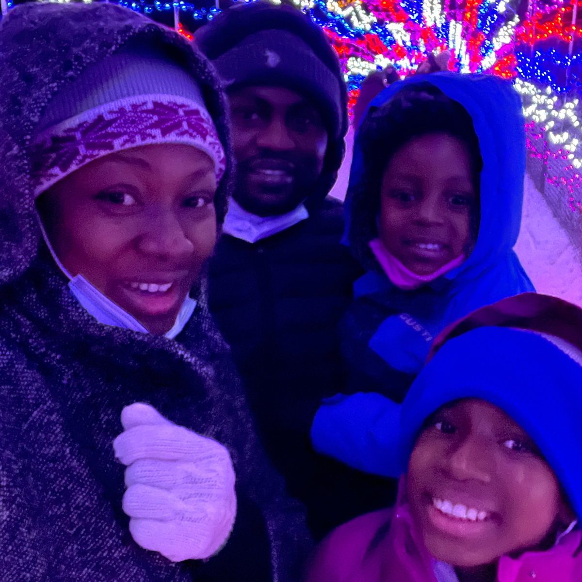 A young Black family stand together smiling, posing in front of a Christmas lights display with winter clothes on outside. 