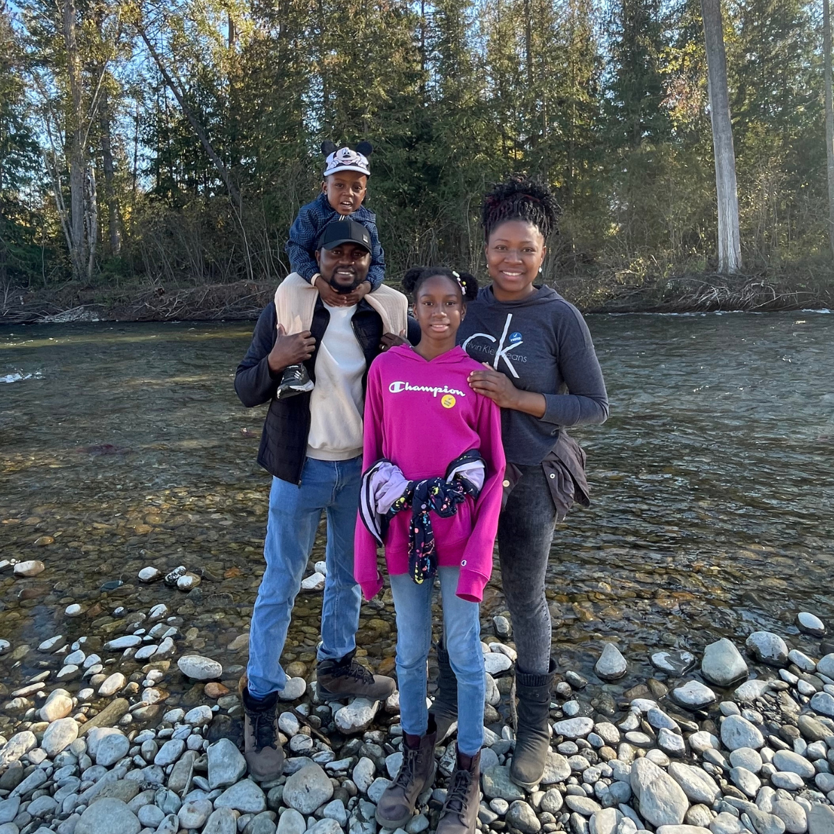 A young Black family stand together smiling, posing in front of an outdoor beautiful stream with trees in the background. 
