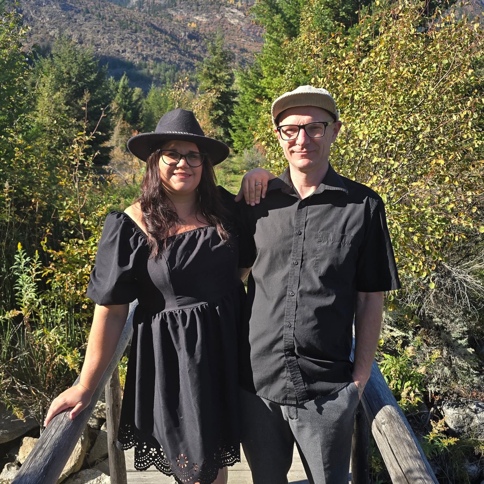 A couple stands between two wooden rails in front of trees. Both are wearing hats, eyeglasses and black clothes.