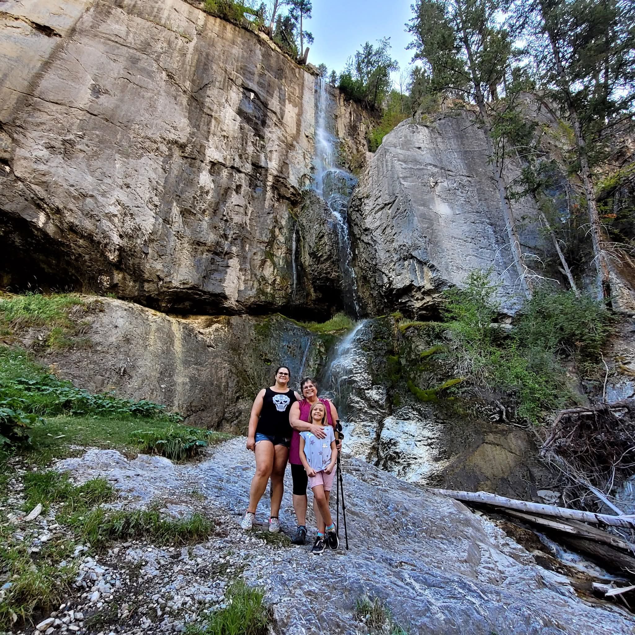 Two adults with a child standing at the base of a mountain, with a rock face waterfall behind them.