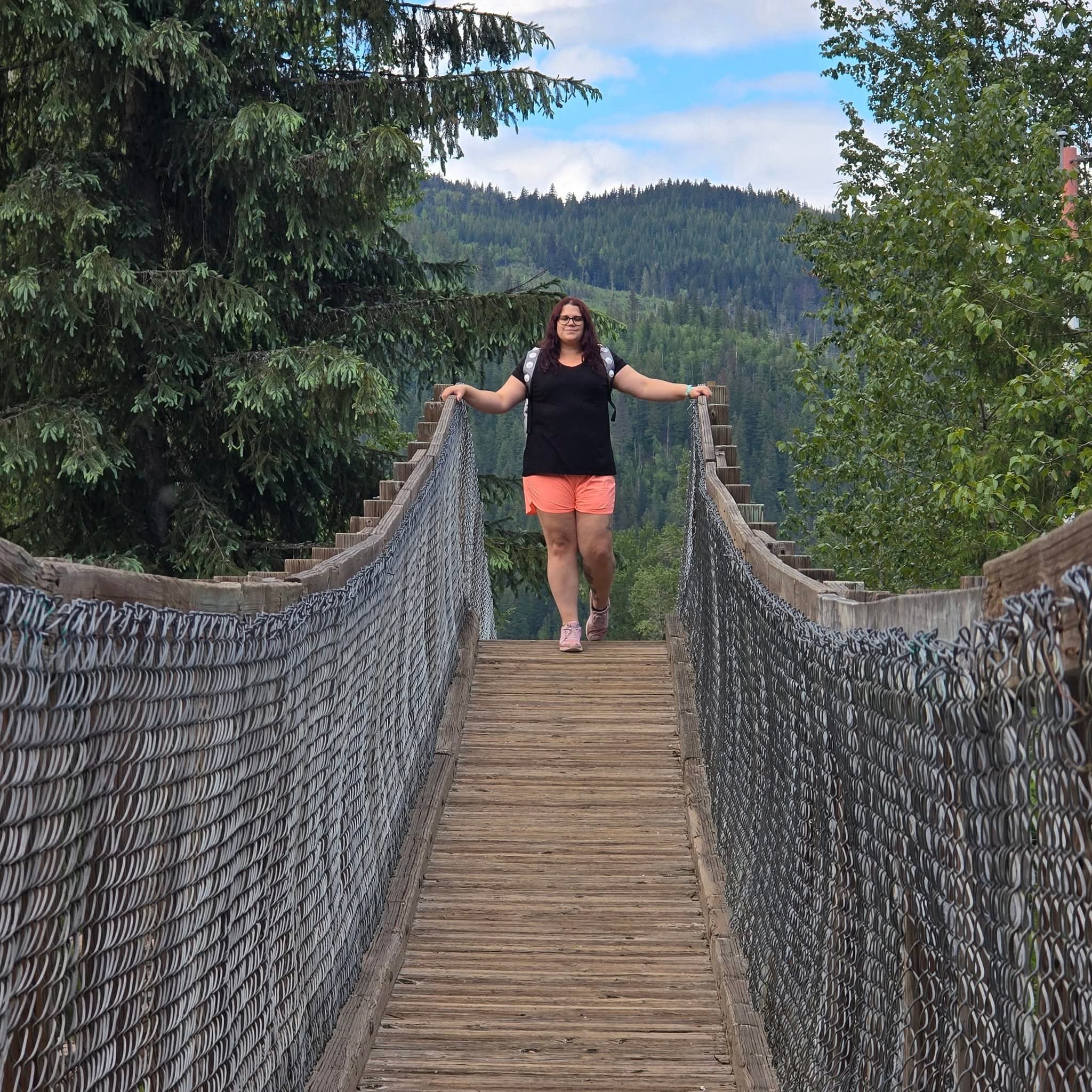 Person standing on a wooden suspension bridge with chain-link sides, surrounded by dense green trees and forested hills under a partly cloudy sky.