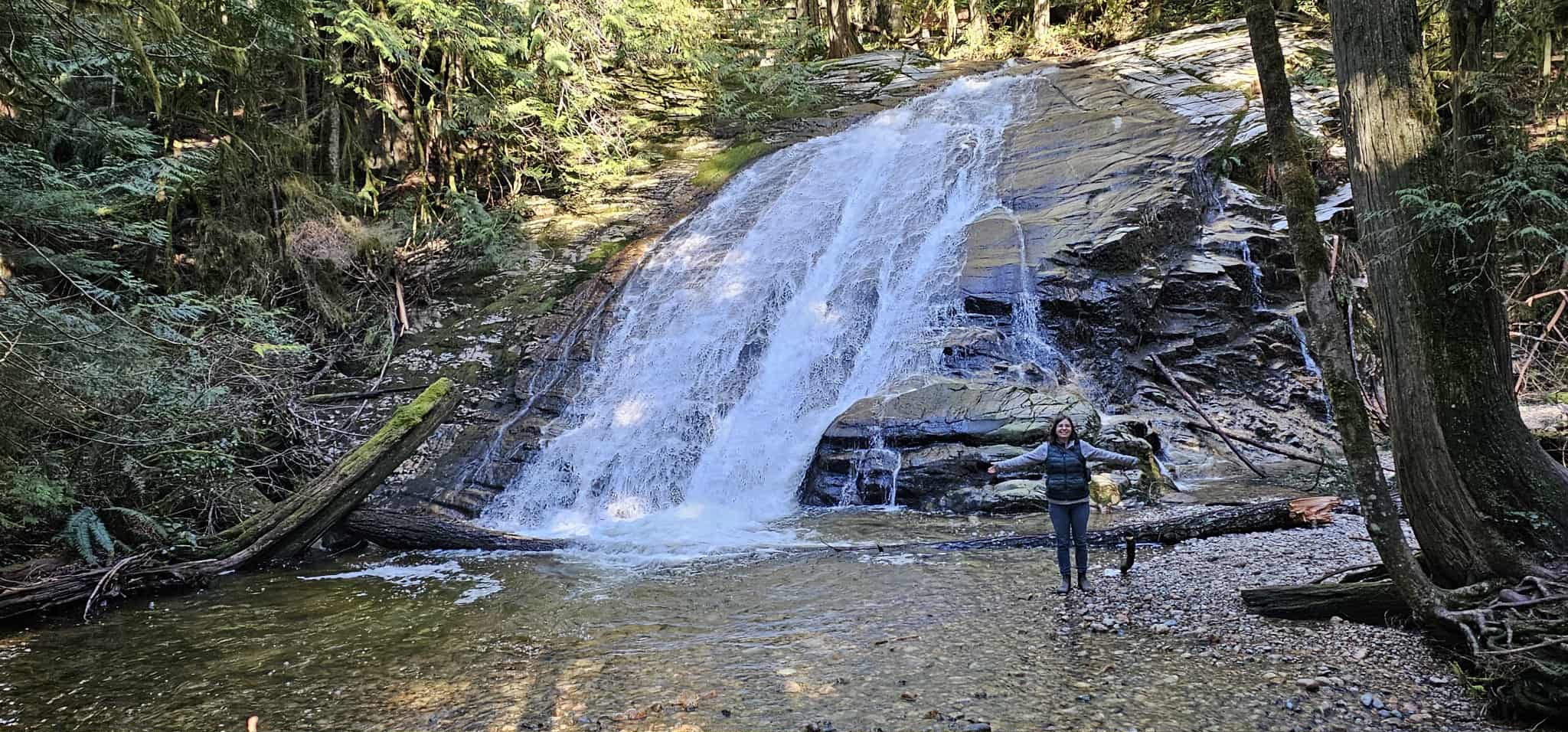 A woman stands at the base of a water falls arms stretched outwards.