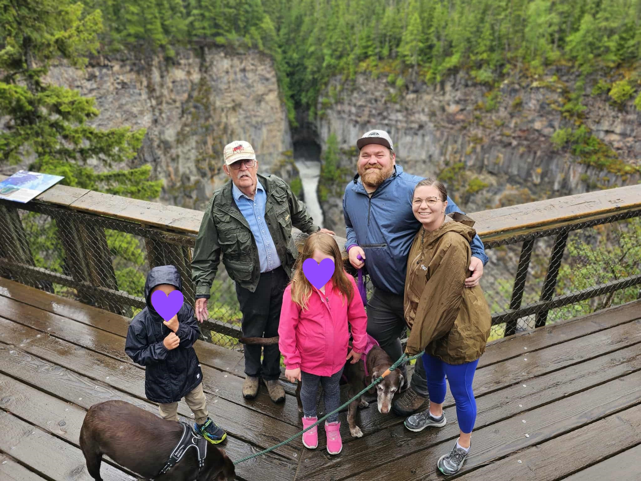 A family of five poses on a walkway overlooking a mountain canyon and river includes an elderly gentleman standing behind 2 children (one wearing a blue jacket, the other a pink jacket) and beside a man and woman with their black dog on a harness and leash.