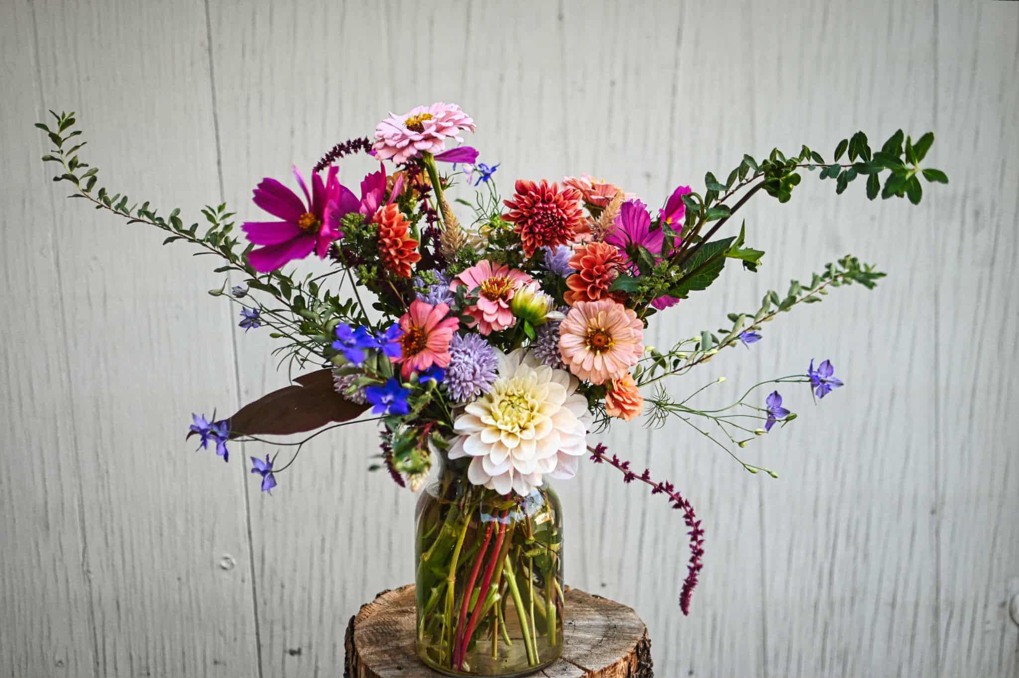 An arrangement of white, pink, blue and purple flowers beautifully arranged in a glass container in front of a white wood panelled wall. 