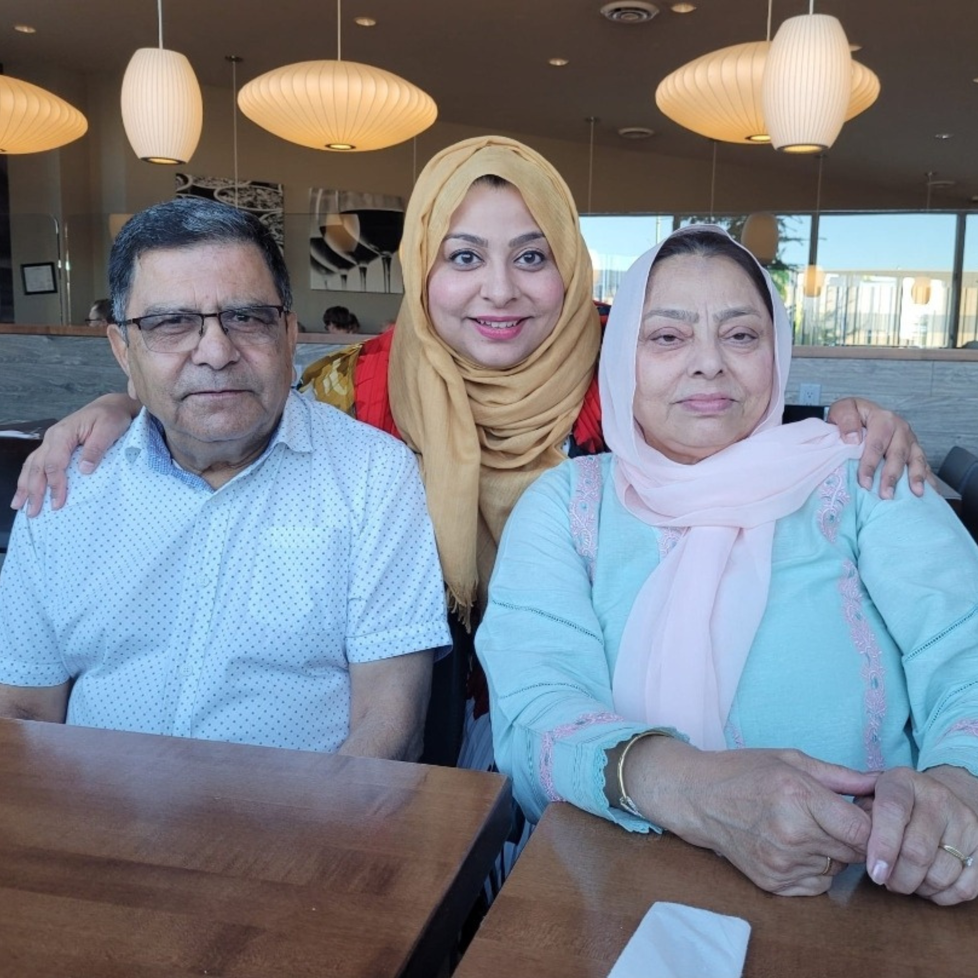 An adult woman standing in the middle of and behind her parents who are sitting at a table, is leaning forward with both hands resting on their shoulders.  