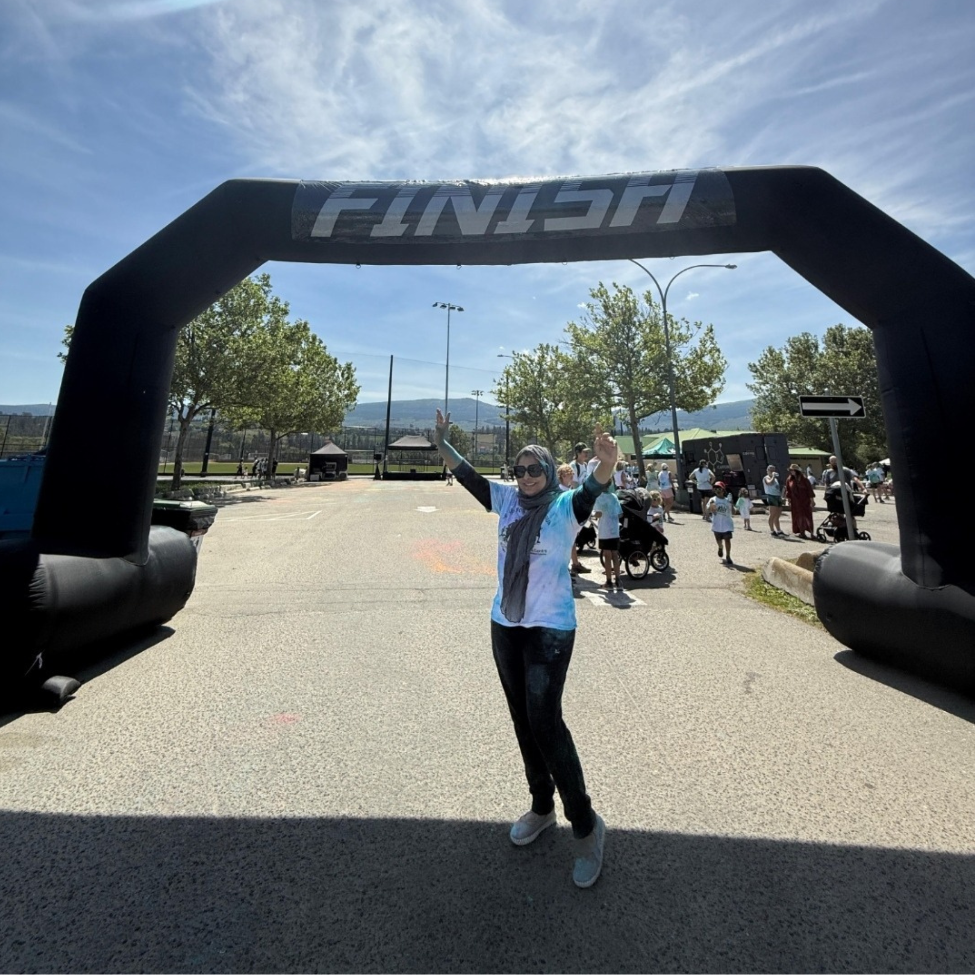 A very bright sunny day, a woman wearing a headscarf, black pants, a white t-shirt over a black long sleeve shirt with her arms raised in a celebratory fashion is standing underneath a black inflatable FINISH sign. 