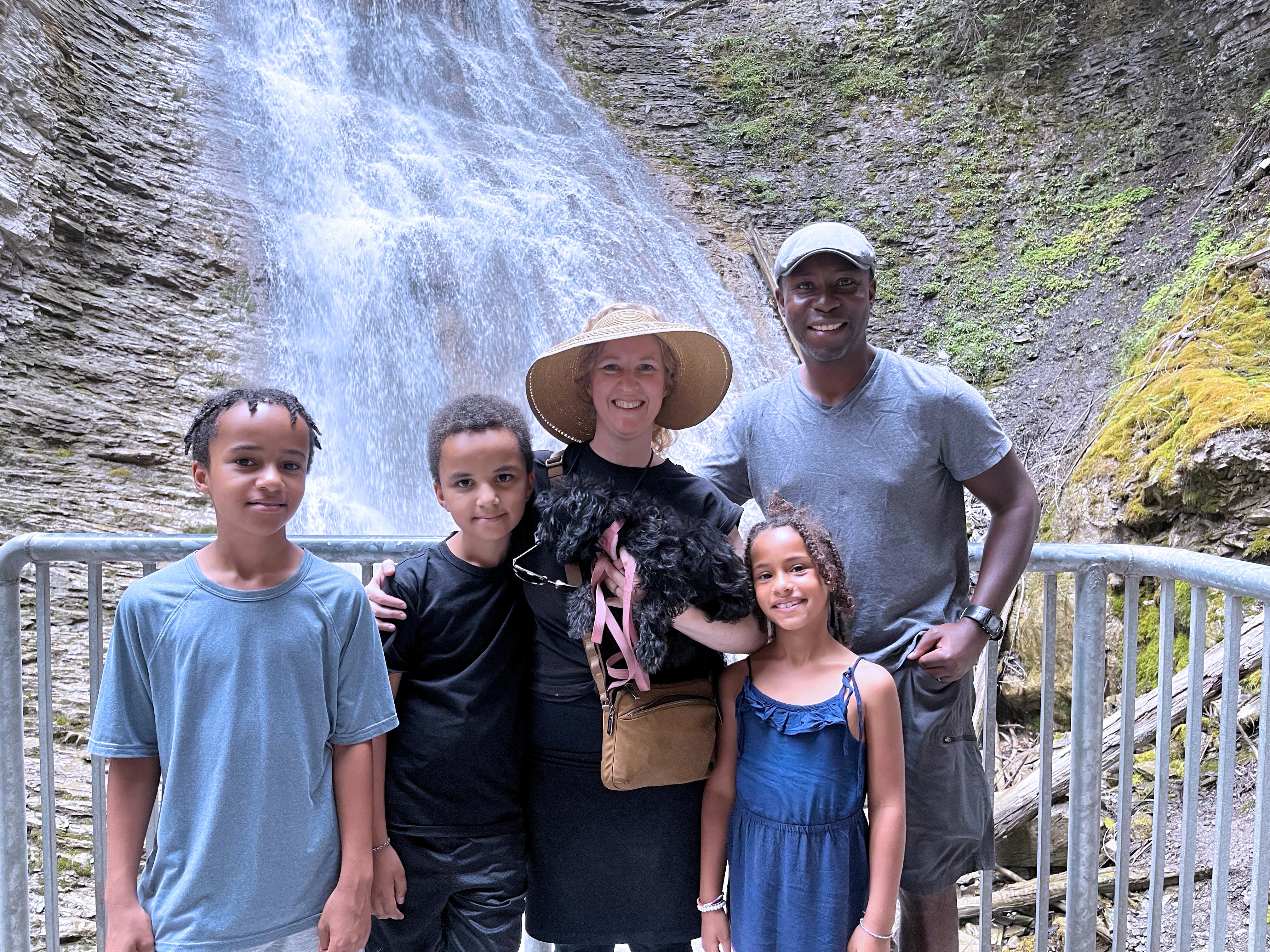 A man stands with two boys, one girl, and a woman all smiling, in front of a waterfall.