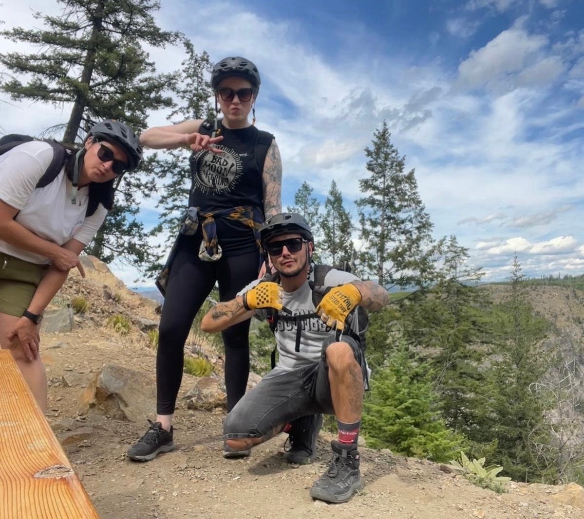 Two women and one man posing with helmets on a mountain in front of trees.