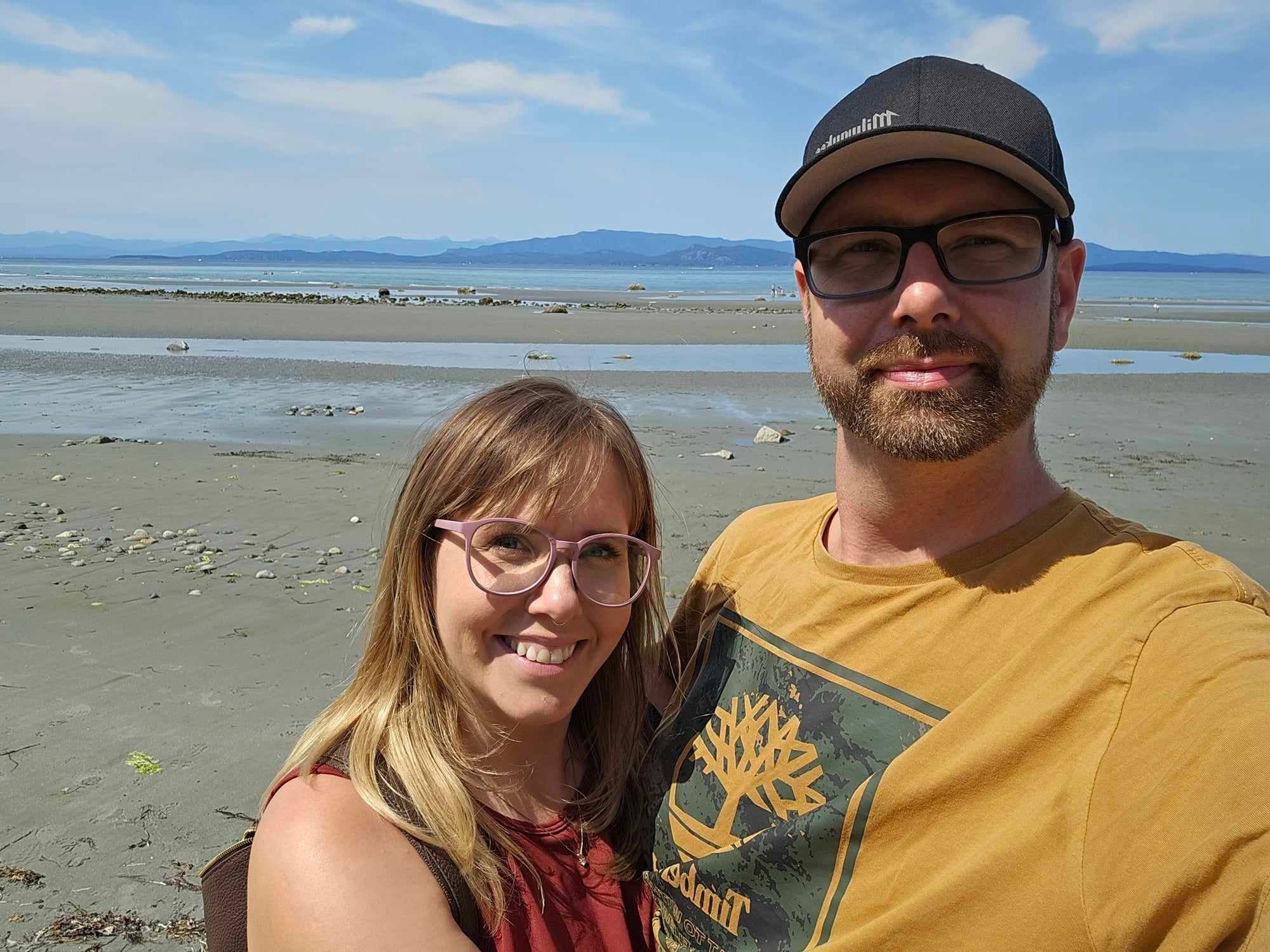 Couple standing on a sandy beach with scattered rocks and a calm sea under a partly cloudy sky.