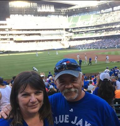 A woman with shoulder-length brown hair stands next to a man with grey hair, a baseball cap and a goatee, who has his arm around her. They are both wearing blue t-shirts and standing in front of a crowd at a baseball game. 